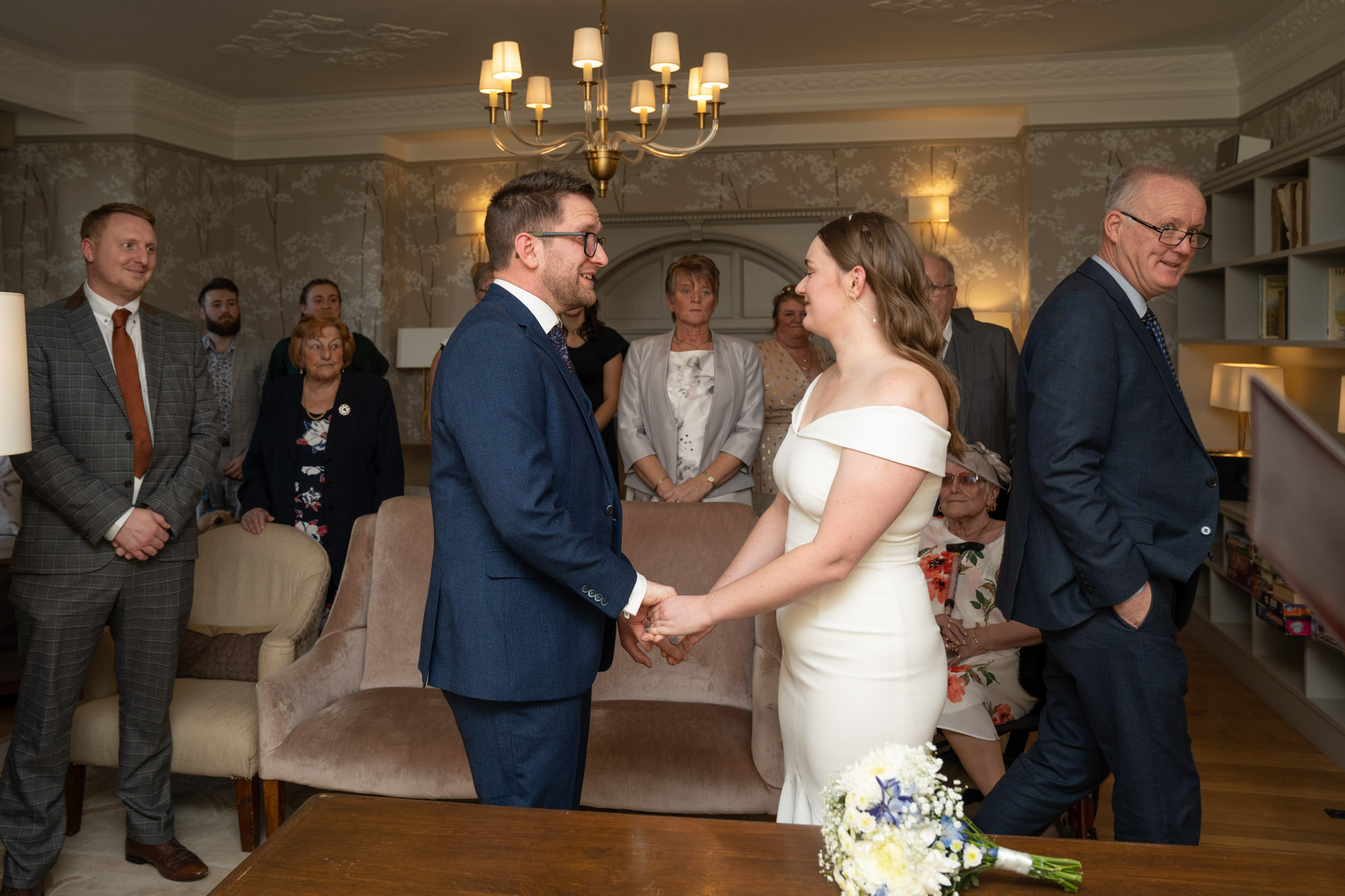 Bride and groom holding hands and looking at each other as the ceremony begins inside Crag Wood, guests standing behind them and her father stepping back to the side