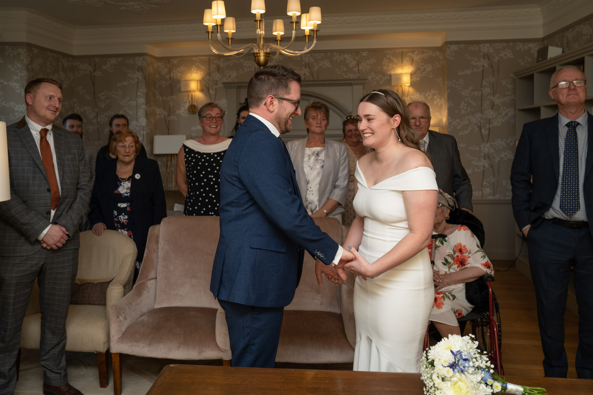 Bride and groom holding hands and smiling at each other at the front of the ceremony room at Crag Wood, family and friends watching on either side