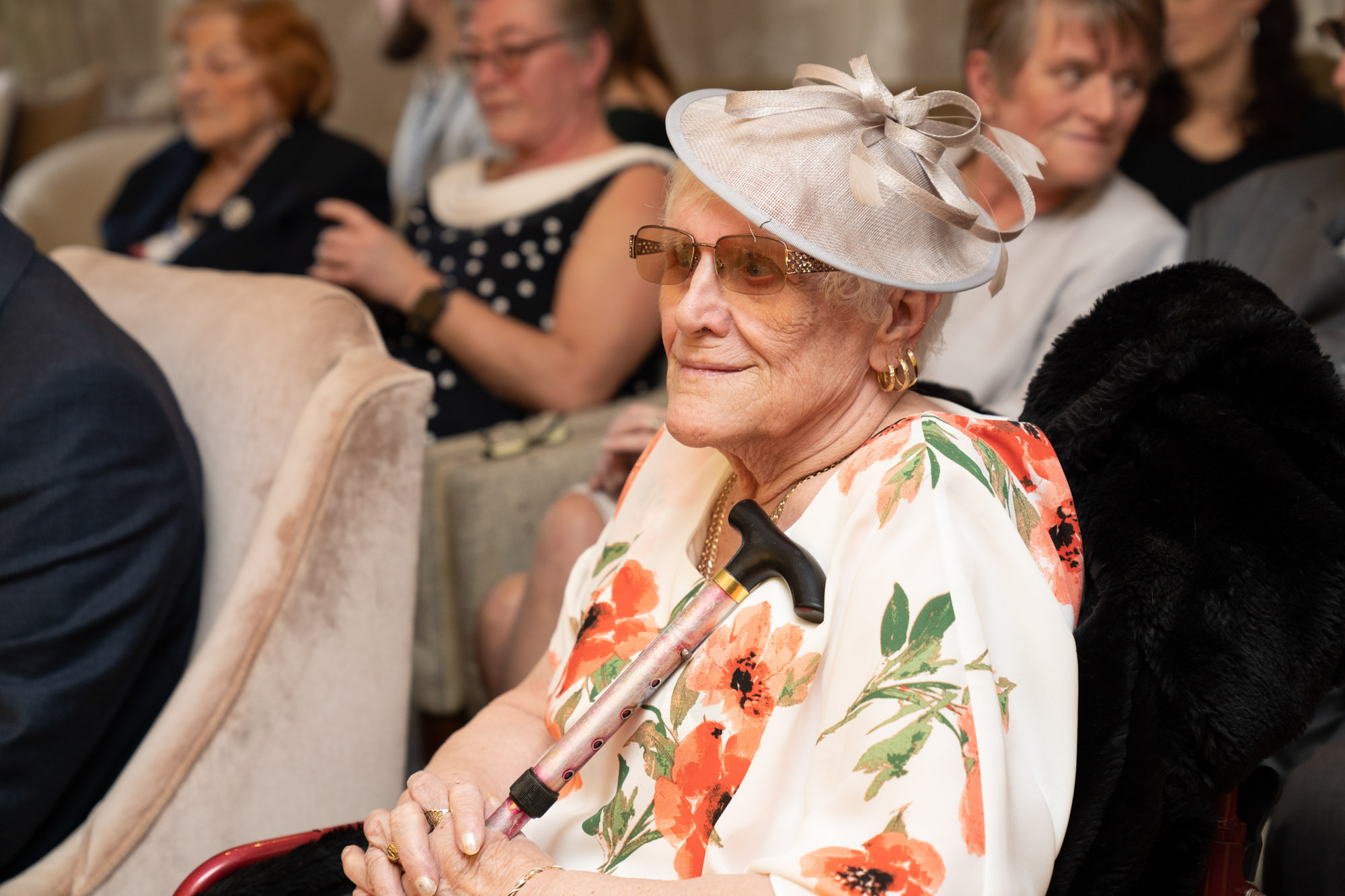 Elderly female wedding guest seated in the ceremony room at Crag Wood, wearing a floral cape and a cream fascinator, smiling softly