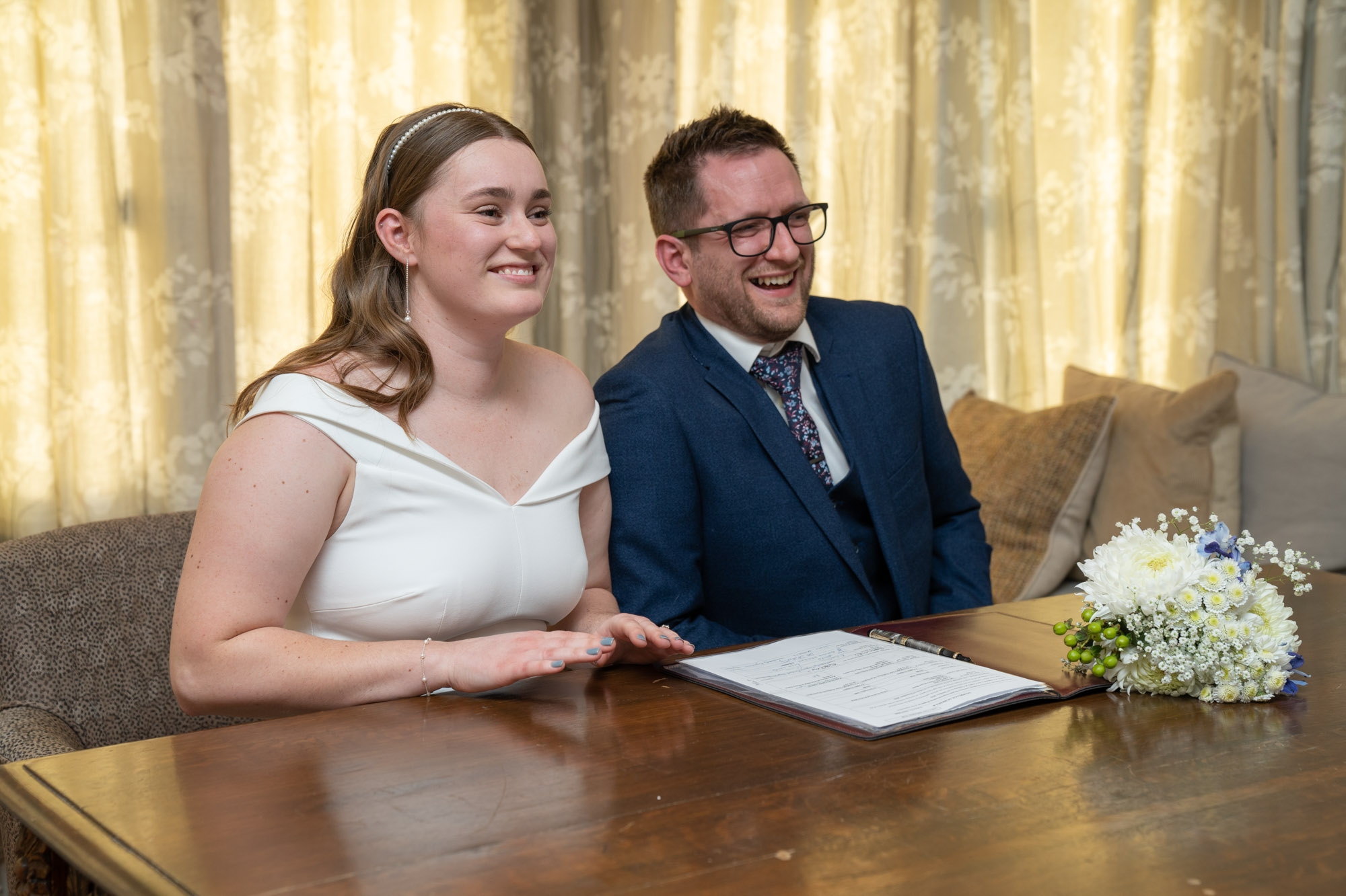 Bride and groom seated at a table laughing together during the register signing, wedding bouquet of white flowers beside the open register