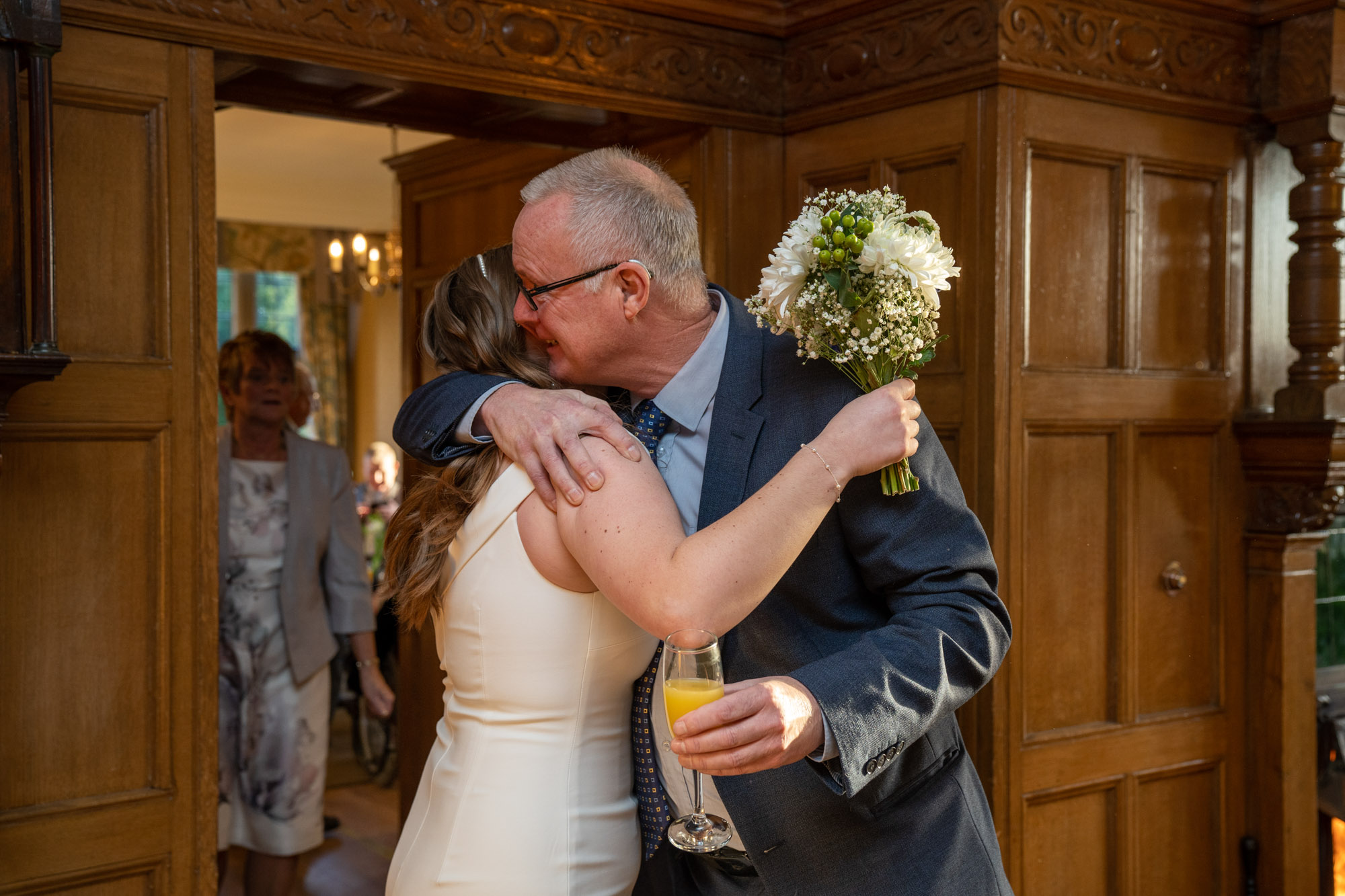 Bride being hugged tightly by an older man in a suit inside the panelled hallway at Crag Wood, he is still holding her bouquet and a glass of orange juice