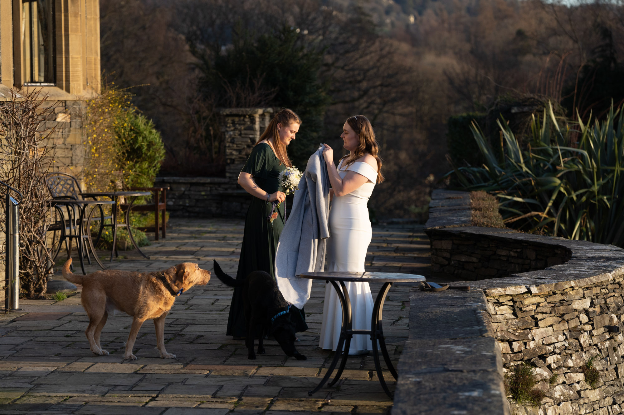 Bride and a woman in a green dress on the stone terrace outside Crag Wood, the bride holding a grey coat over a bistro table while two dogs look on in winter sunshine