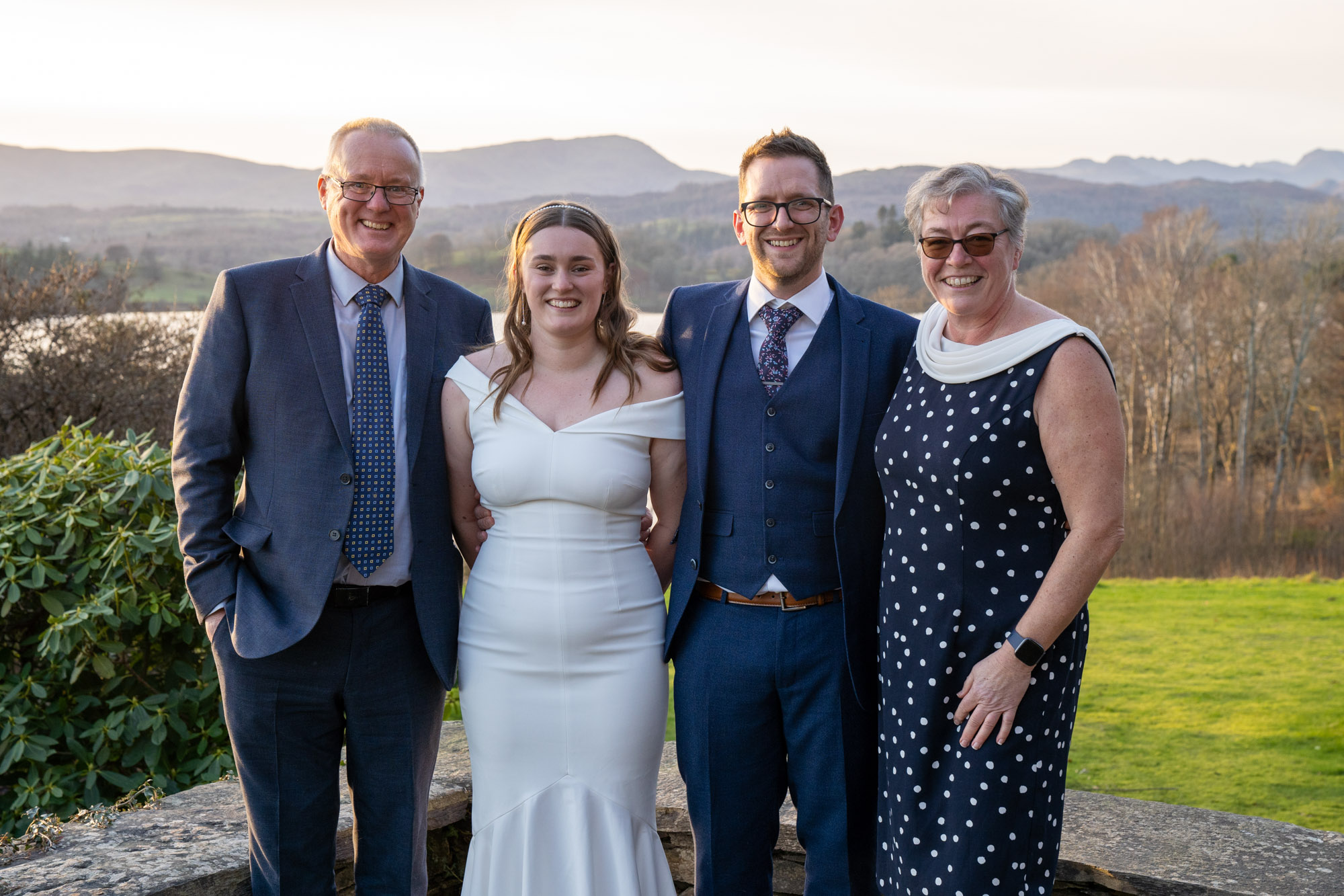 Bride and groom with two older guests, smiling together on the terrace at Crag Wood, Windermere, hills and lake in the background