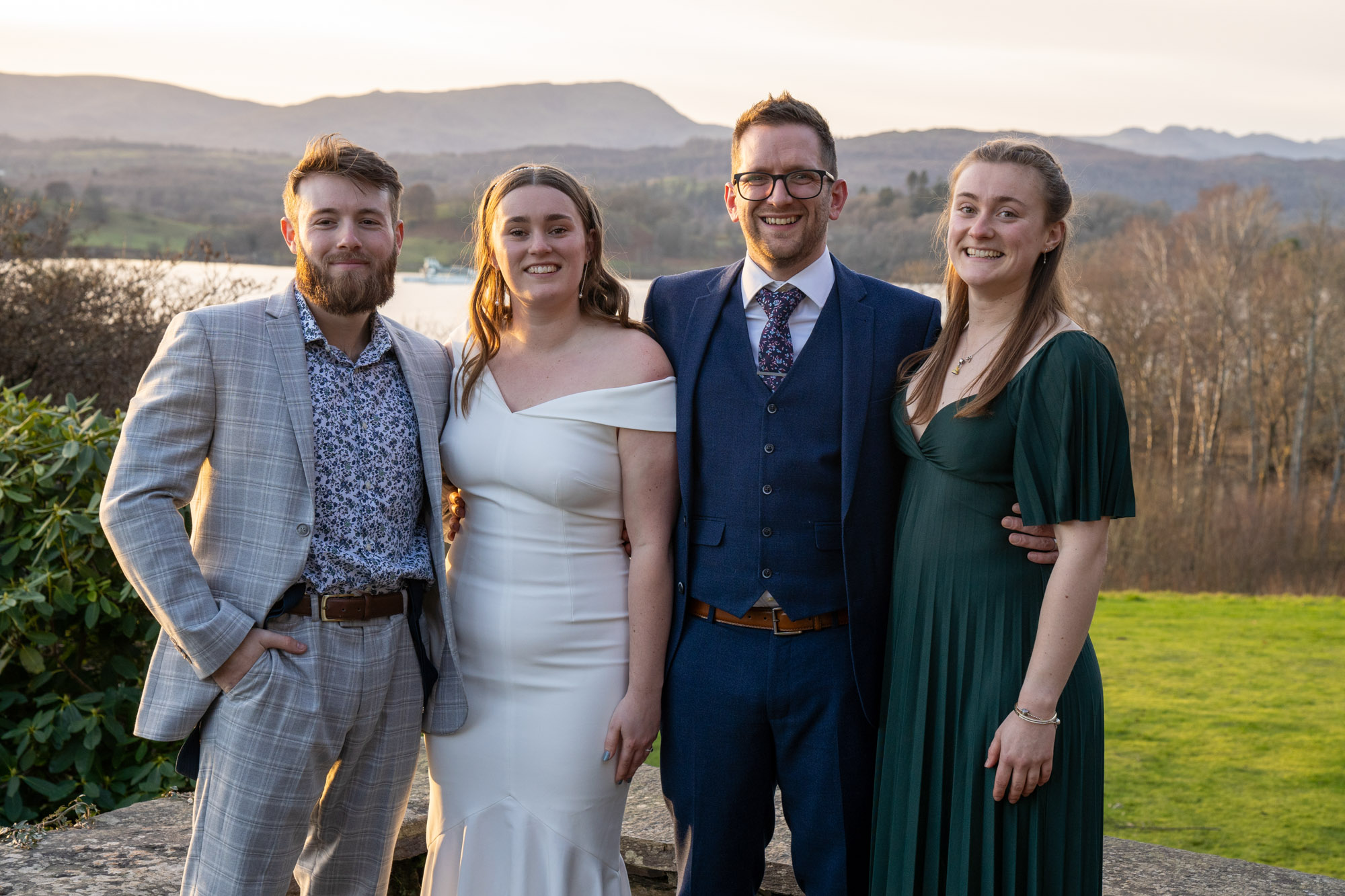 Bride and groom with two guests, all smiling, on the stone terrace at Crag Wood with Lake Windermere and the Cumbrian fells visible behind