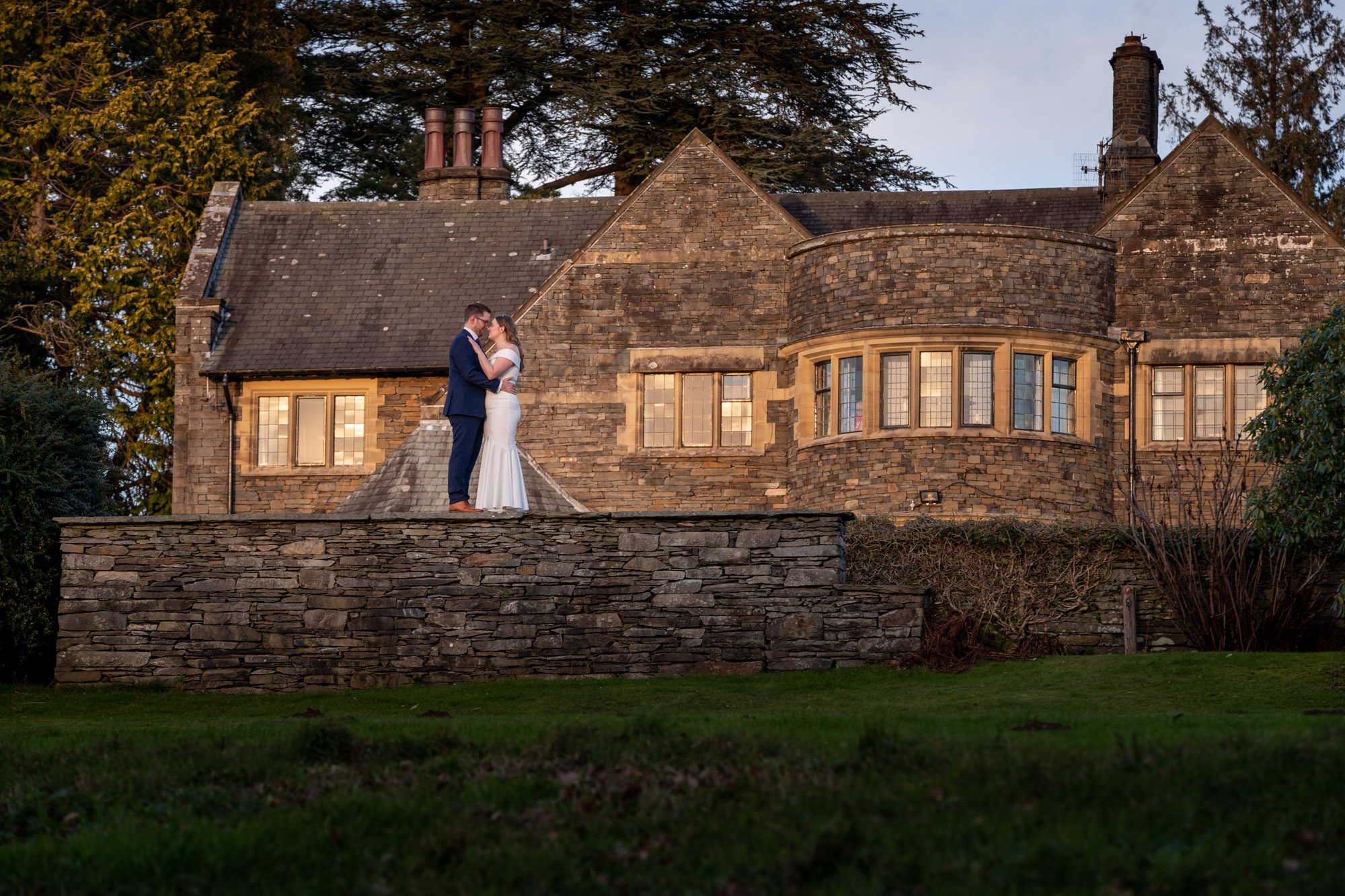 Bride and groom standing close on a low stone wall in front of Crag Wood manor house, bathed in warm golden late-afternoon light
