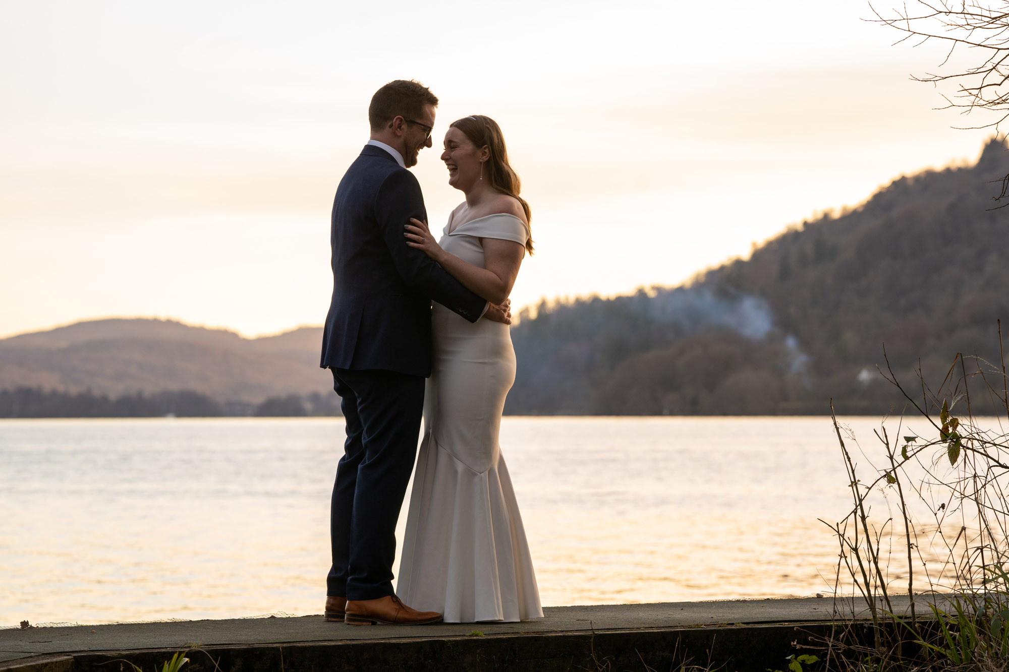 Bride laughing as she looks up at the groom on a jetty at Windermere, hills and pale evening sky reflected in the lake behind them