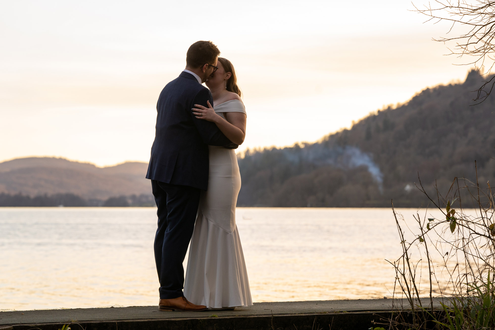 Bride and groom kissing on the Windermere jetty at sunset, the lake and Cumbrian hills glowing amber behind them, bare winter branches framing the right side