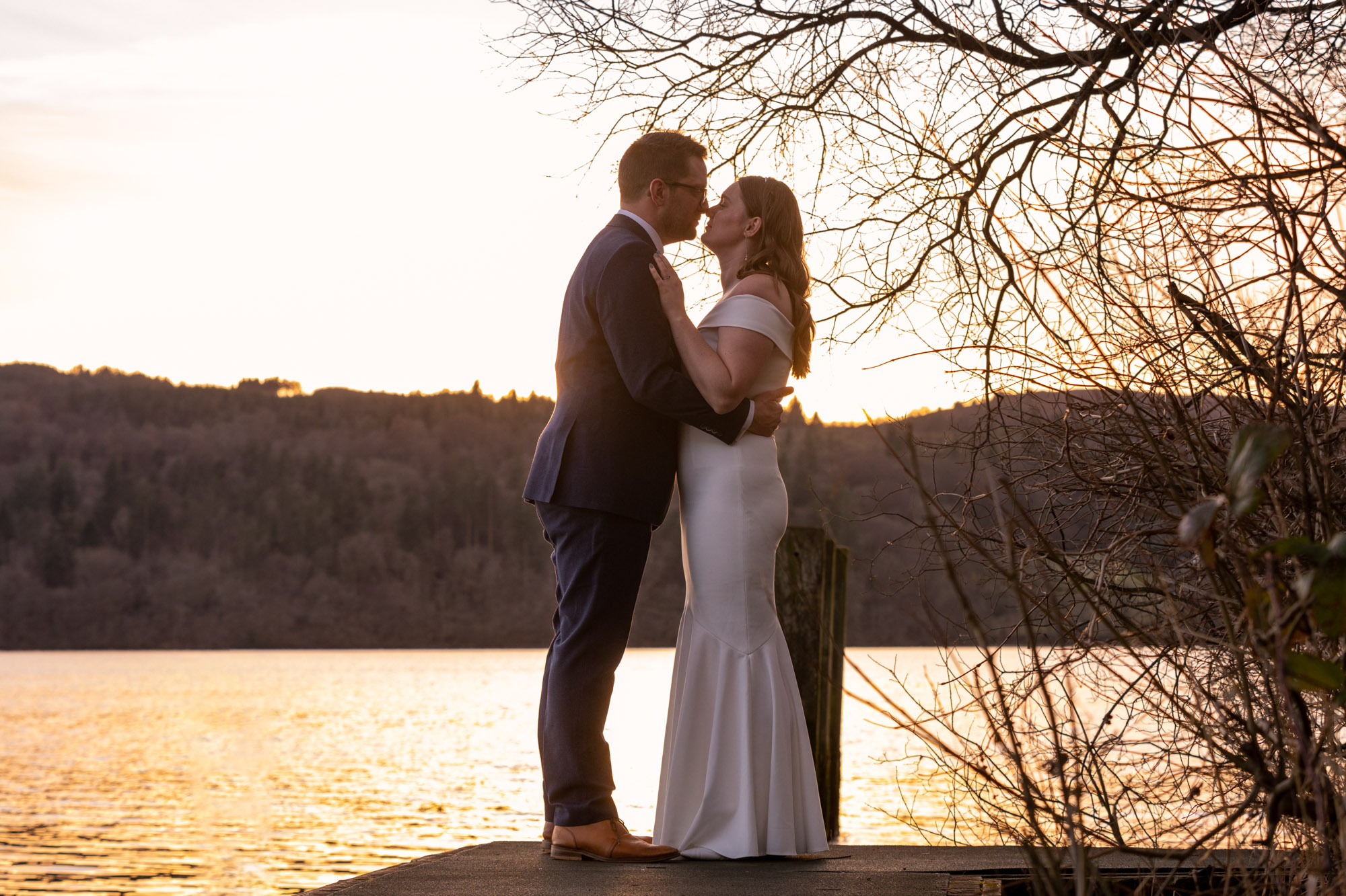 Bride and groom about to kiss on a wooden jetty on Lake Windermere at golden hour, silhouetted against warm orange water and wooded fell behind