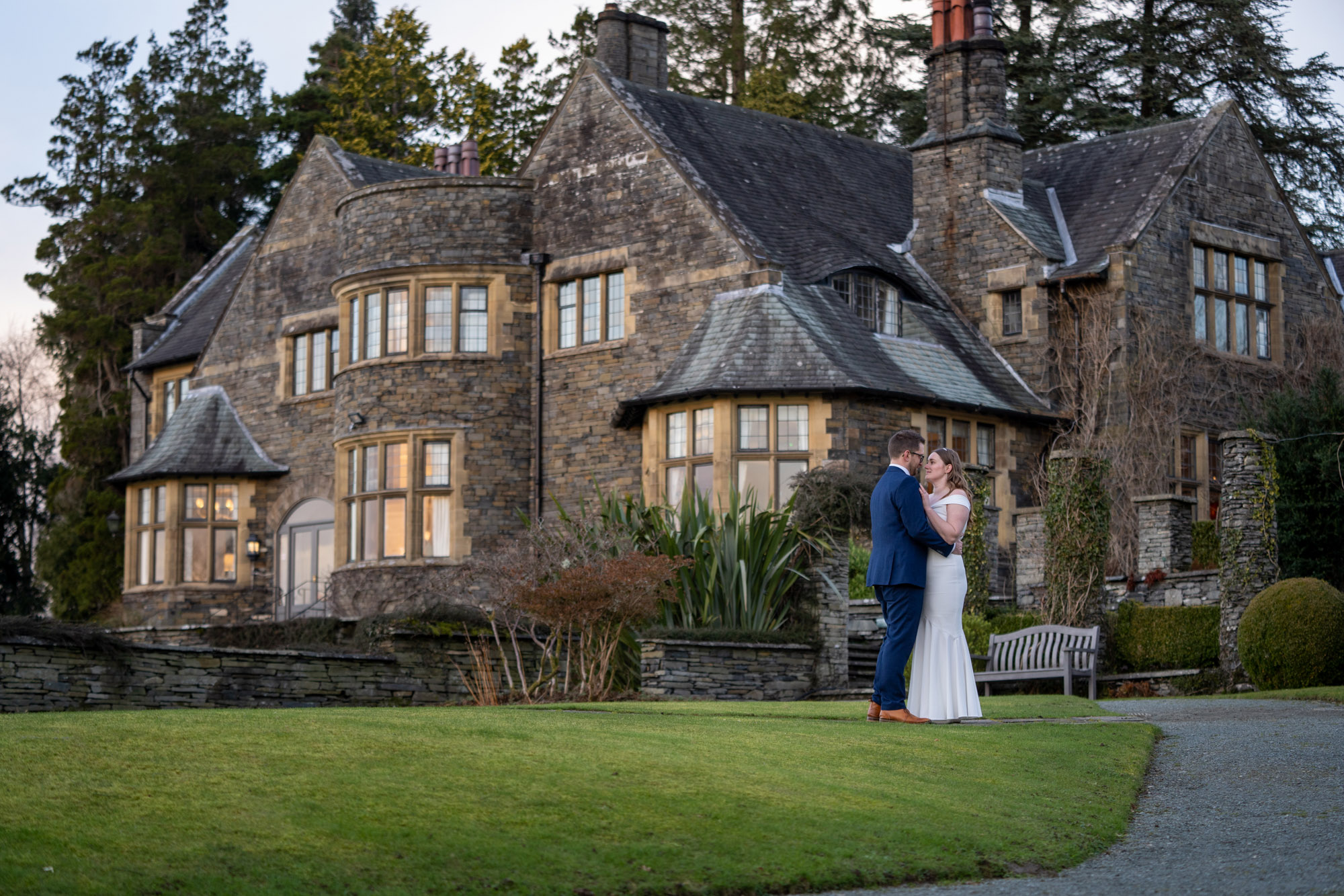 Bride and groom hold each other on the lawn in front of Crag Wood's stone manor house at dusk, the lit windows glowing behind them