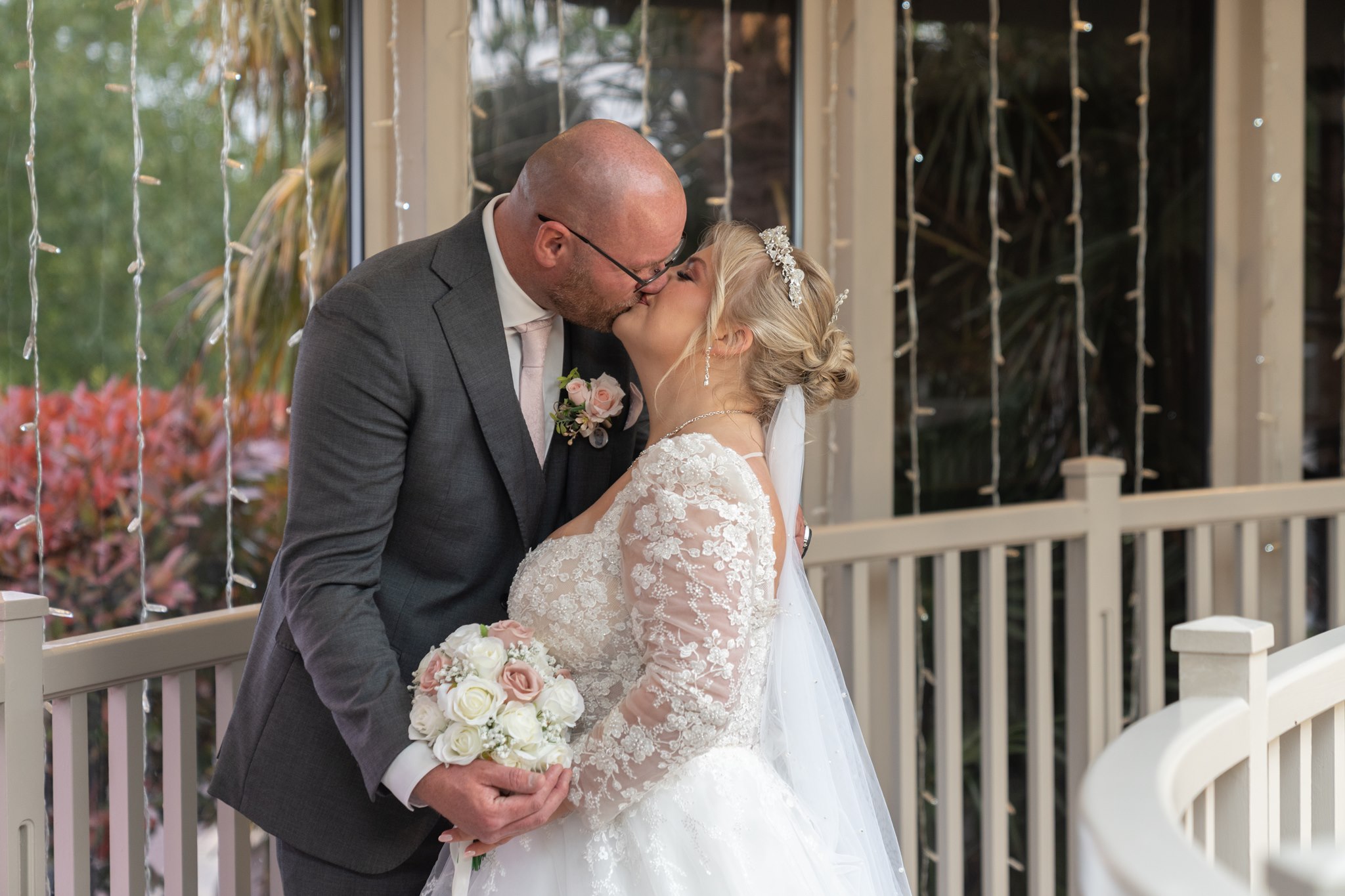 Bride and groom sharing their first kiss on a verandah at Barton Manor, Preston. The bride wears a long-sleeved lace gown and holds a bouquet of white and blush pink roses. The groom wears a grey suit with a pink tie and matching buttonhole. Curtains of fairy lights hang in the background alongside lush green and red garden foliage.