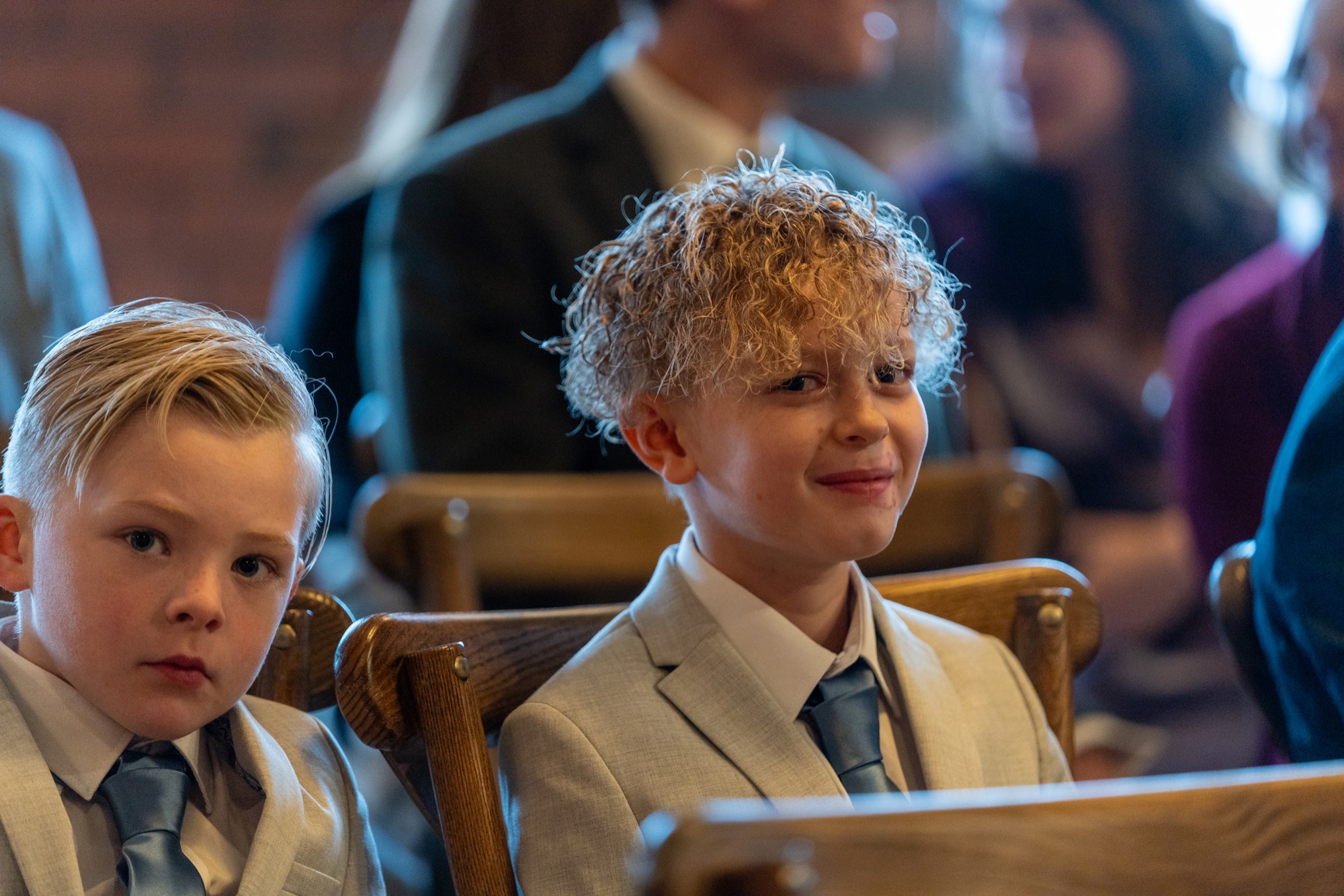 Two young boys in pale suits sitting on wooden chairs waiting for the ceremony to begin