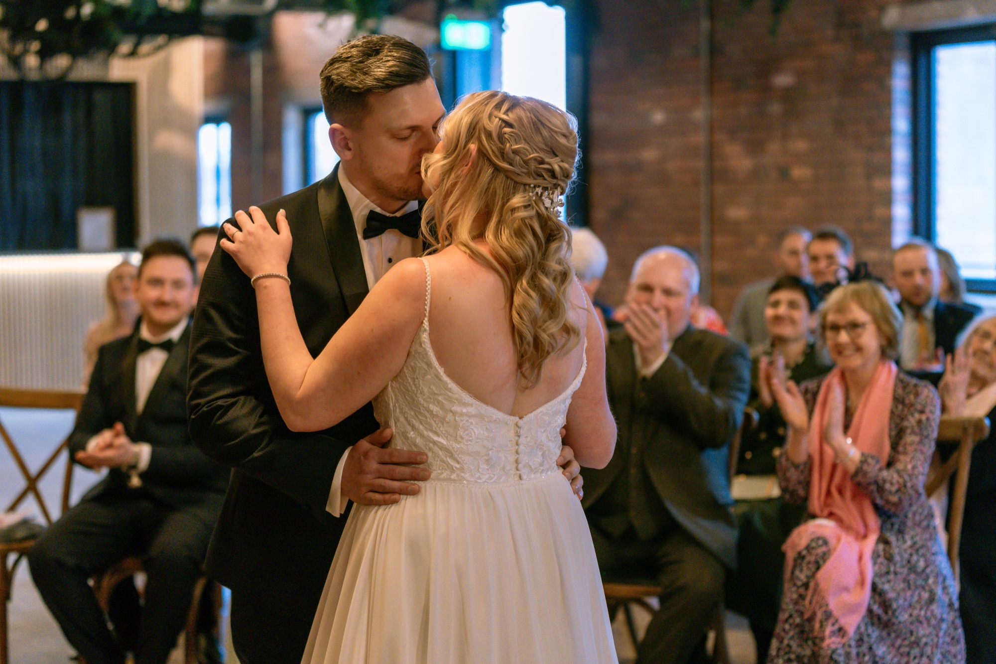 The newlyweds sharing their first kiss as guests applaud in a brick-walled room