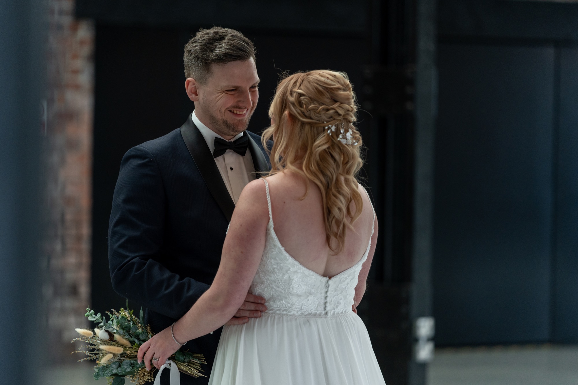 The groom smiling at the bride while holding her bouquet, her back to the camera