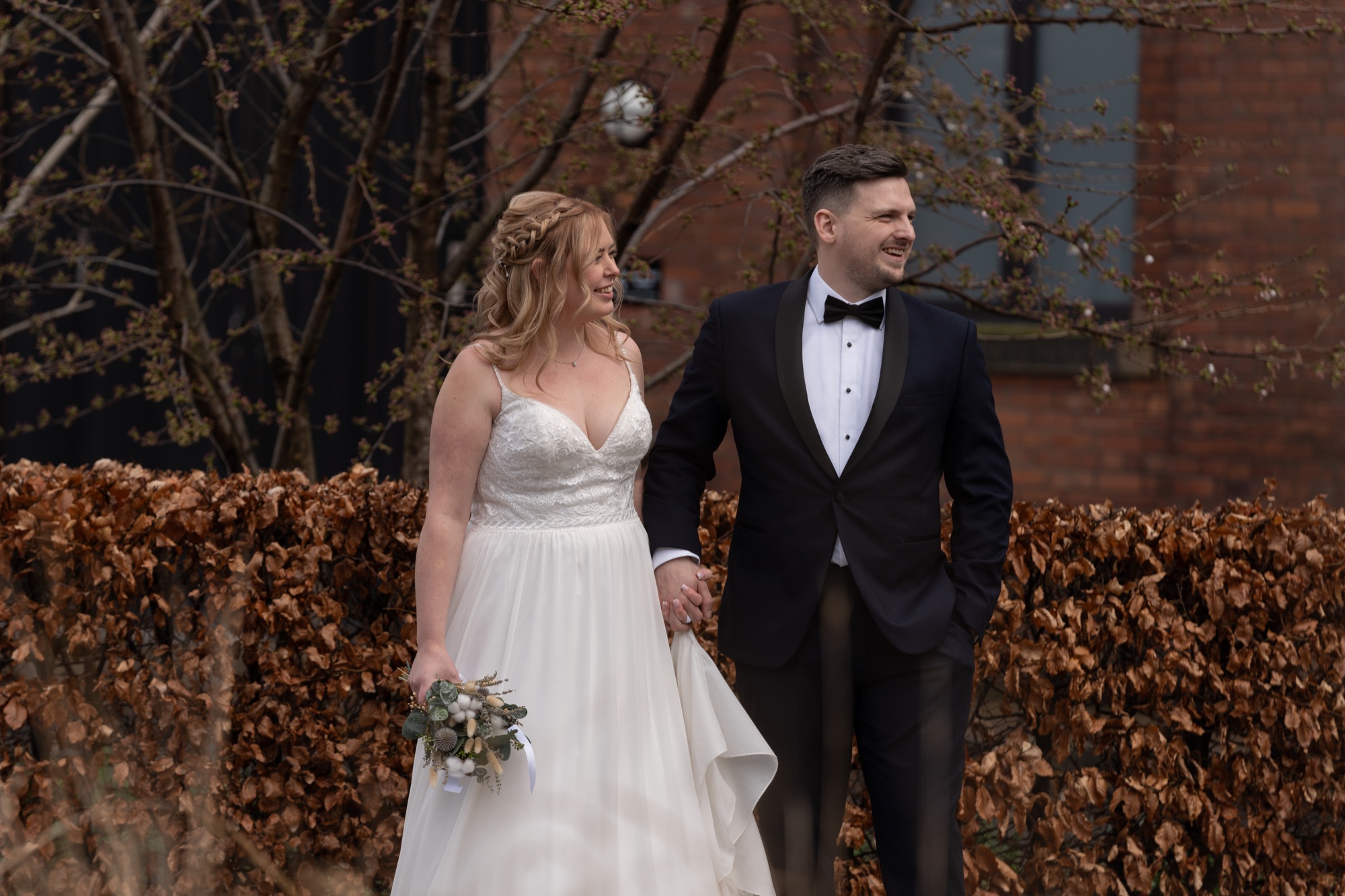The bride and groom walking hand in hand past a copper-leaved hedge and bare winter trees