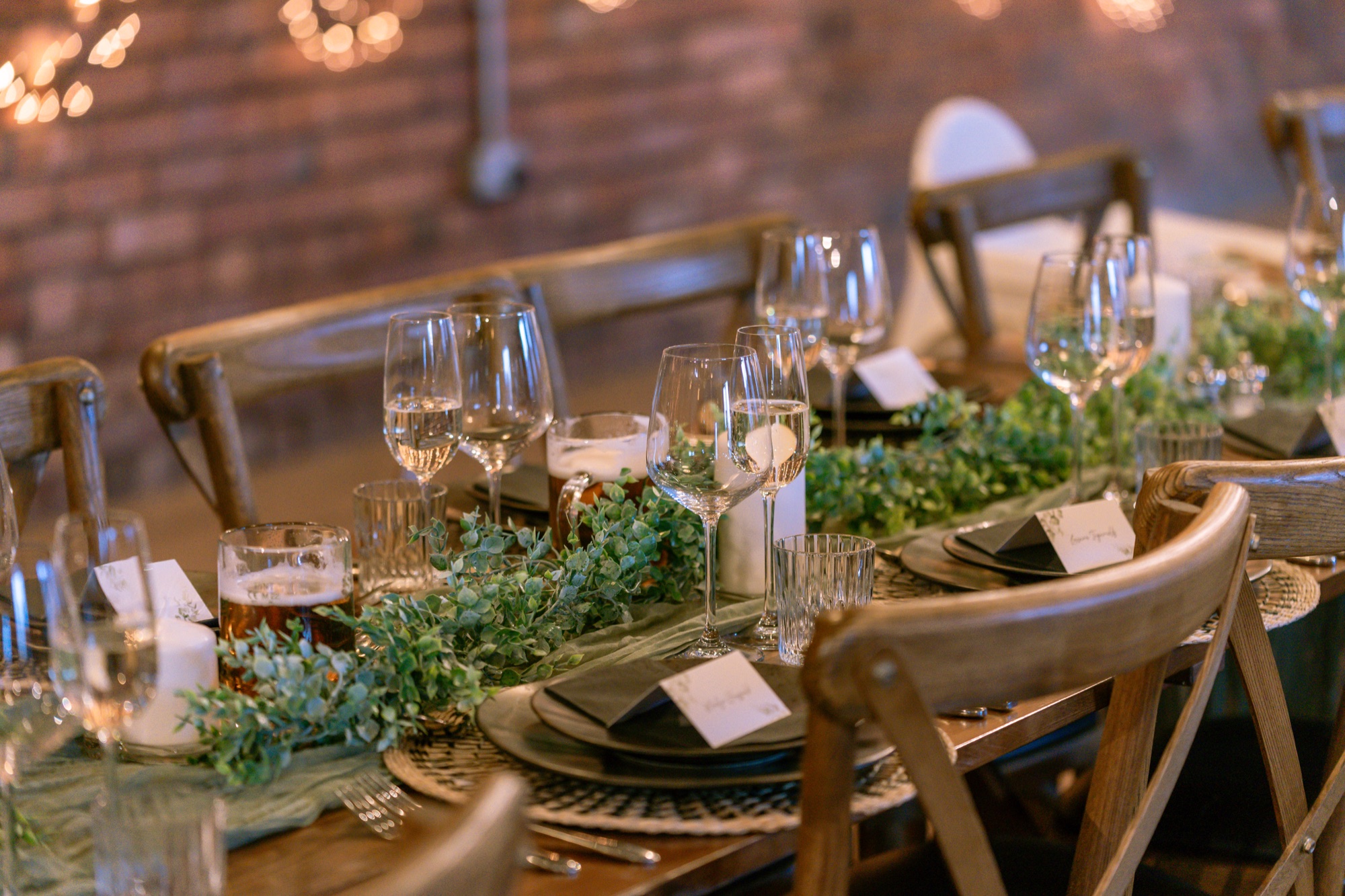 A reception table set with wine glasses, greenery runners and place cards against a brick wall with fairy lights