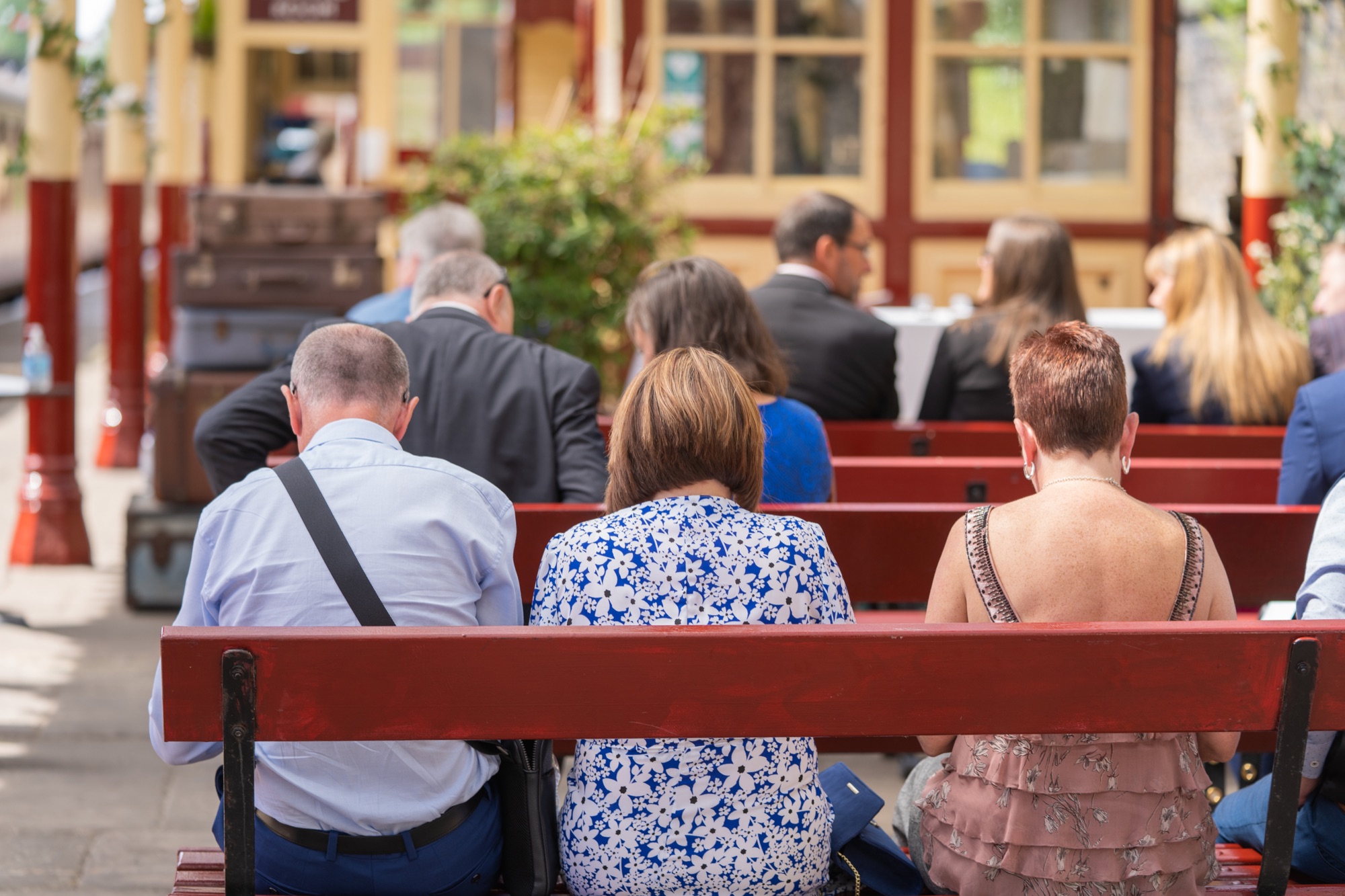Wedding guests sitting with their backs to the camera on red station benches under the Victorian canopy at Bury Bolton Street, waiting for the ceremony to begin