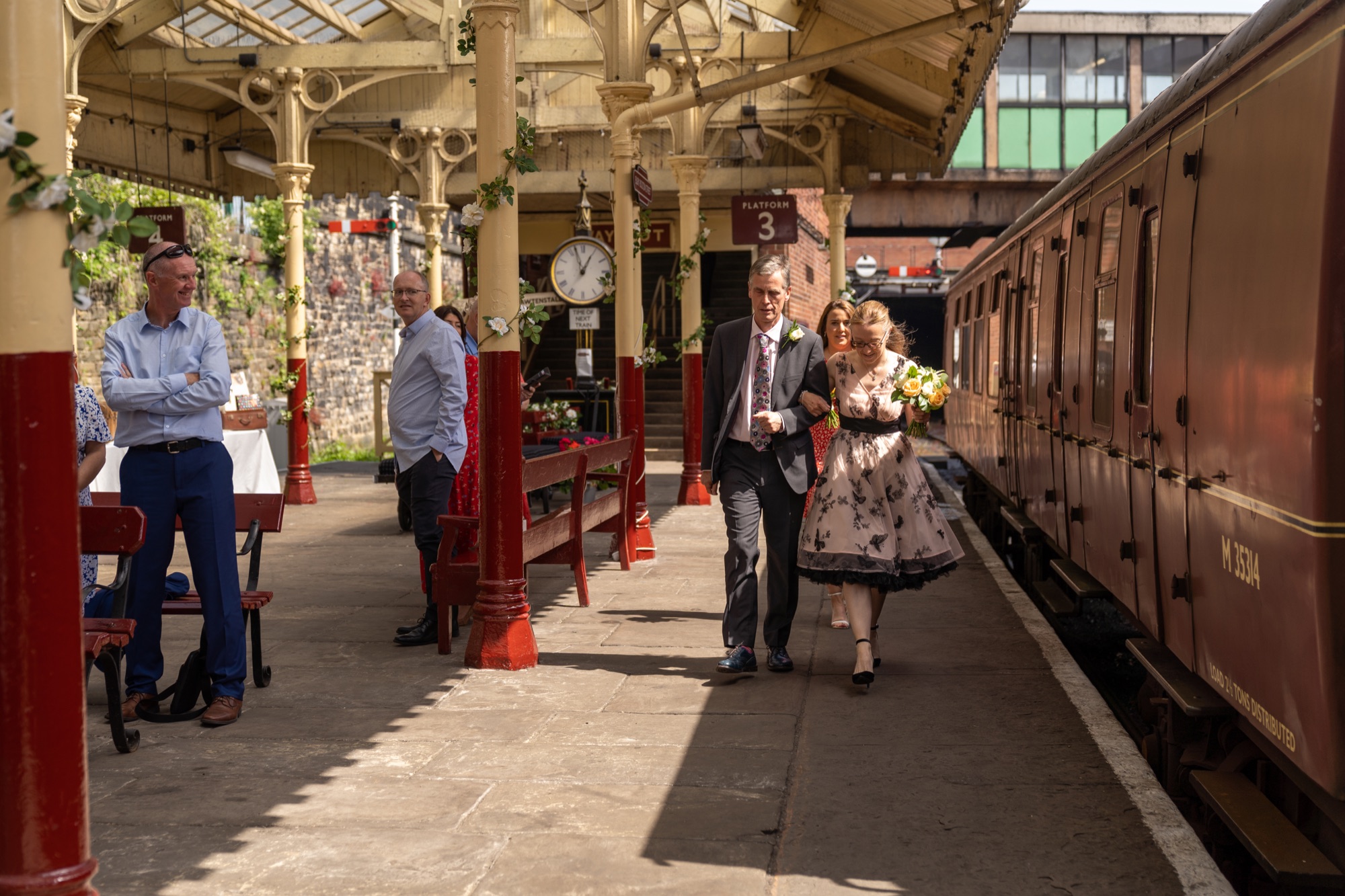 Bride in a floral dress walking along the platform with her father past a maroon heritage carriage and steam locomotive