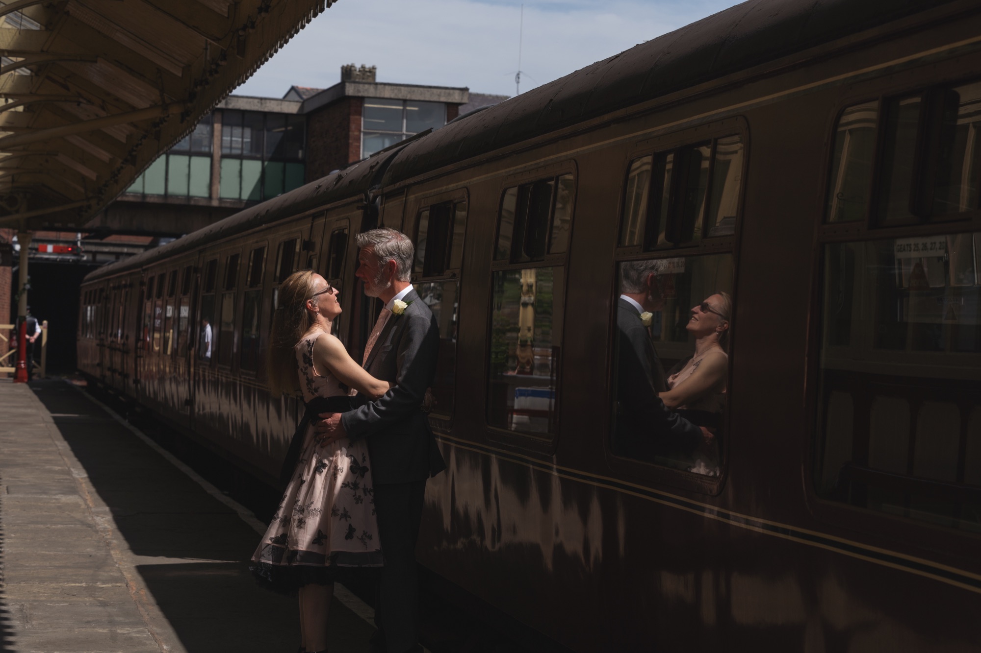 Bride and groom kissing on the platform beside a maroon heritage carriage, reflections in the carriage windows