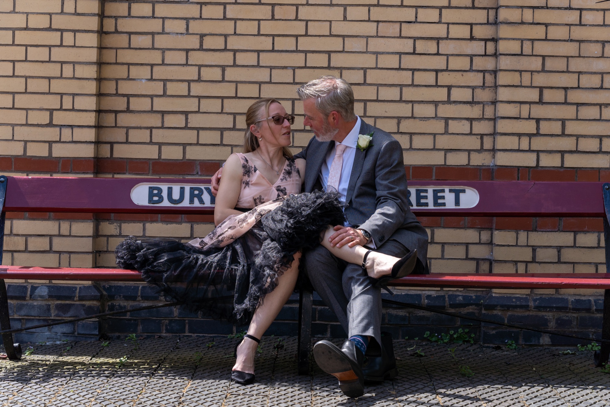 Bride and groom embracing closely on the station bench beneath the Bury Bolton Street sign
