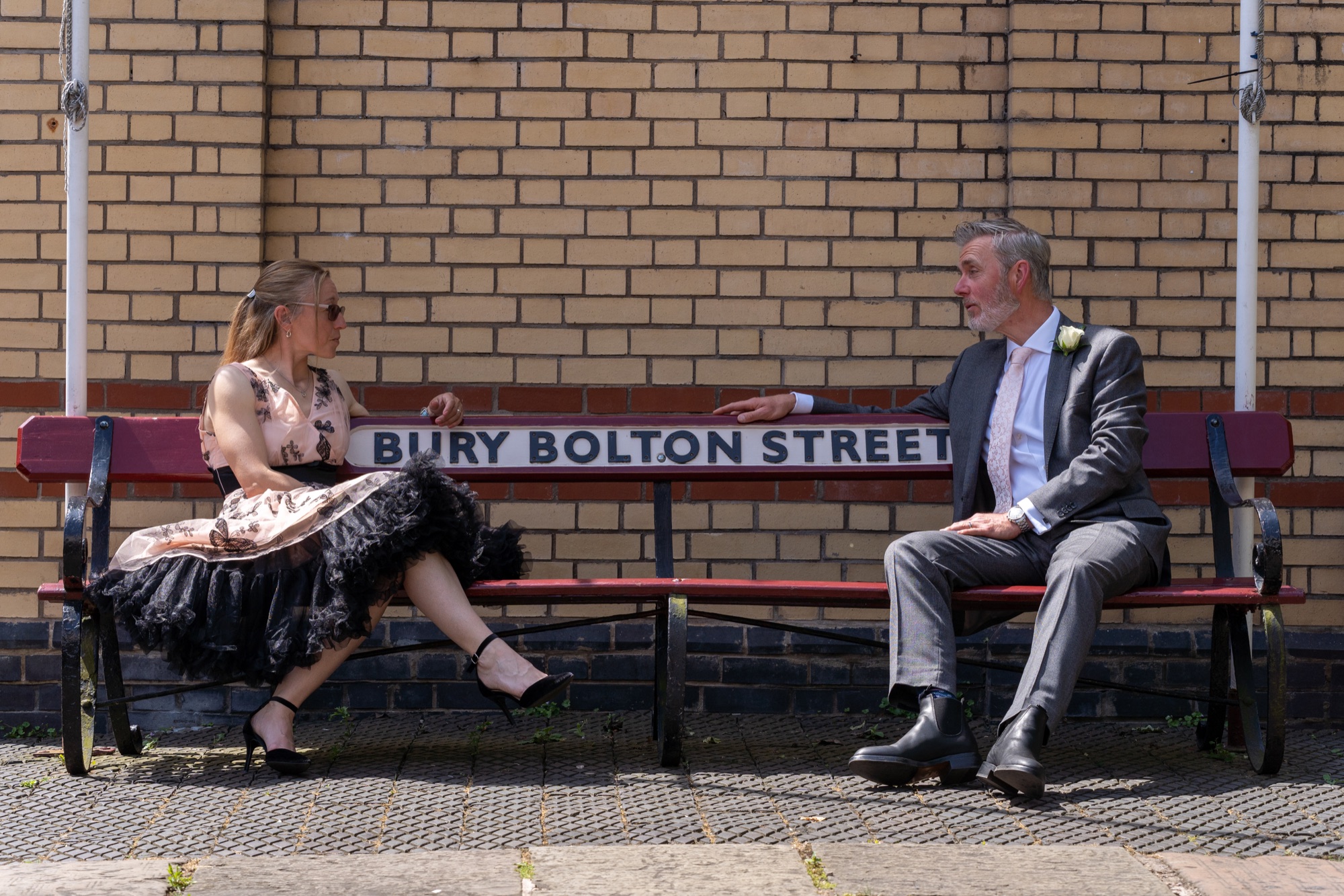 Bride and groom sitting and talking on a red station bench beneath the Bury Bolton Street sign