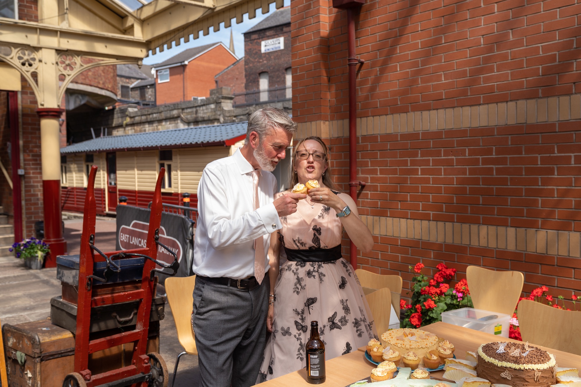 Bride and groom tasting wedding cake together at an outdoor table by the station building