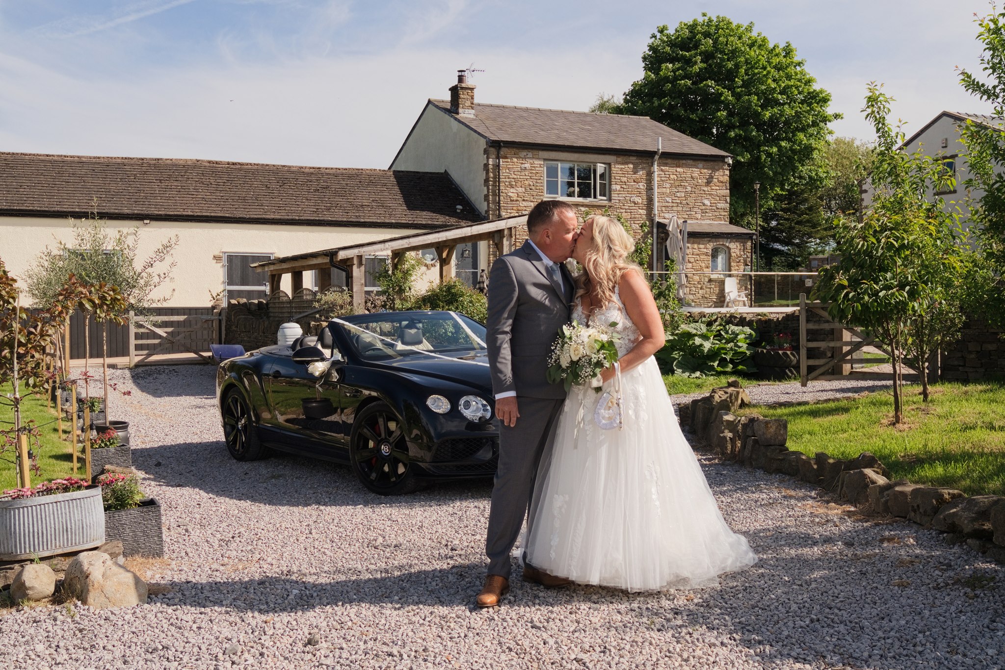 Bride and groom kissing on the gravel driveway of Langshawhead Farm, Darwen, with a black Bentley convertible behind them and the stone farmhouse in the background on a sunny summer day.