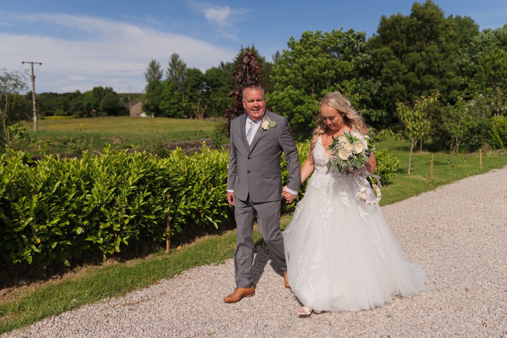 Bride and groom walking hand in hand along the gravel path at Langshawhead Farm, Darwen, with green hedgerows and Lancashire countryside visible behind them under a blue summer sky.