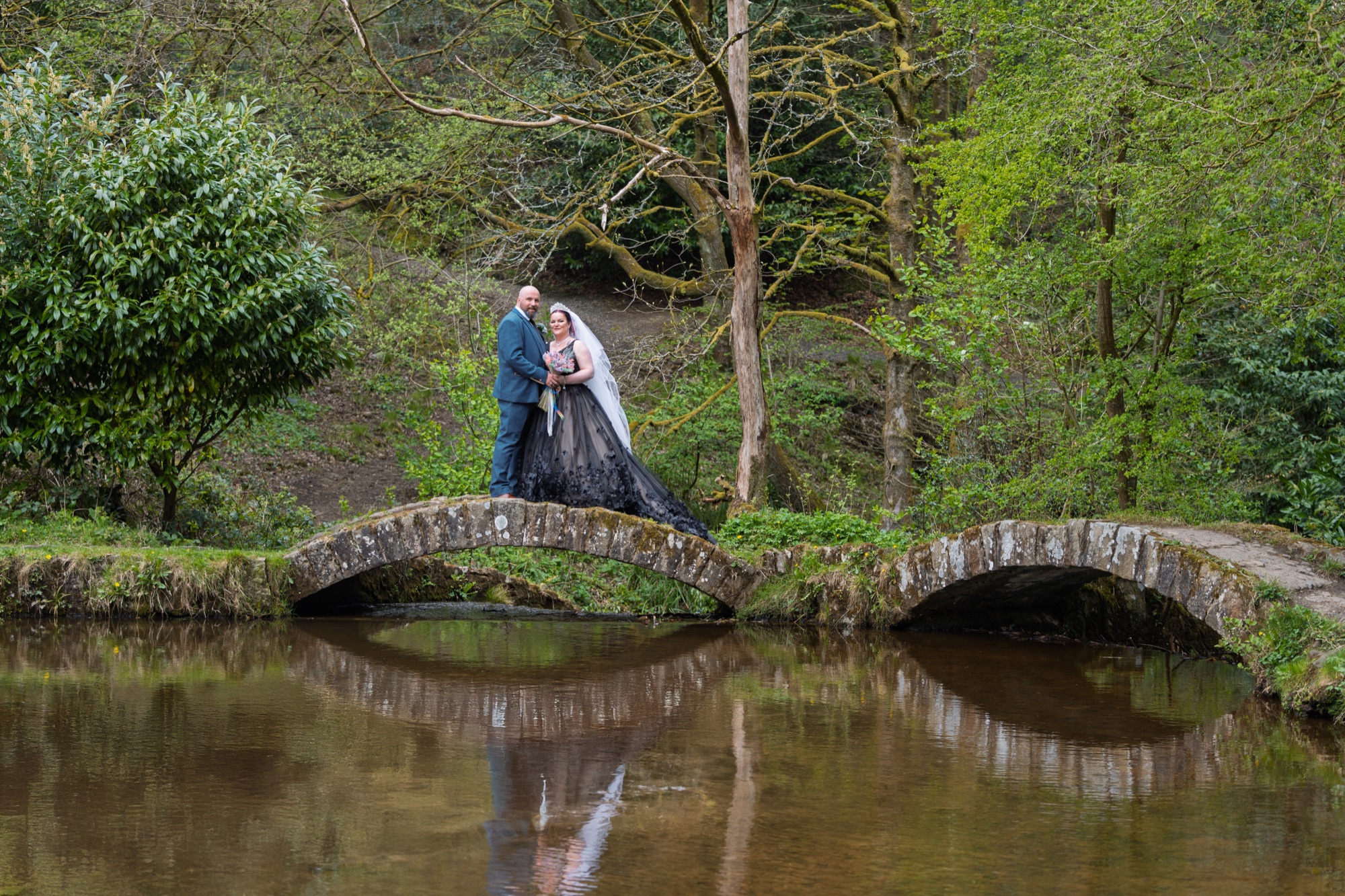 The bride and groom stand together on an old stone bridge over the water in Sunnyhurst Wood, their reflection below.