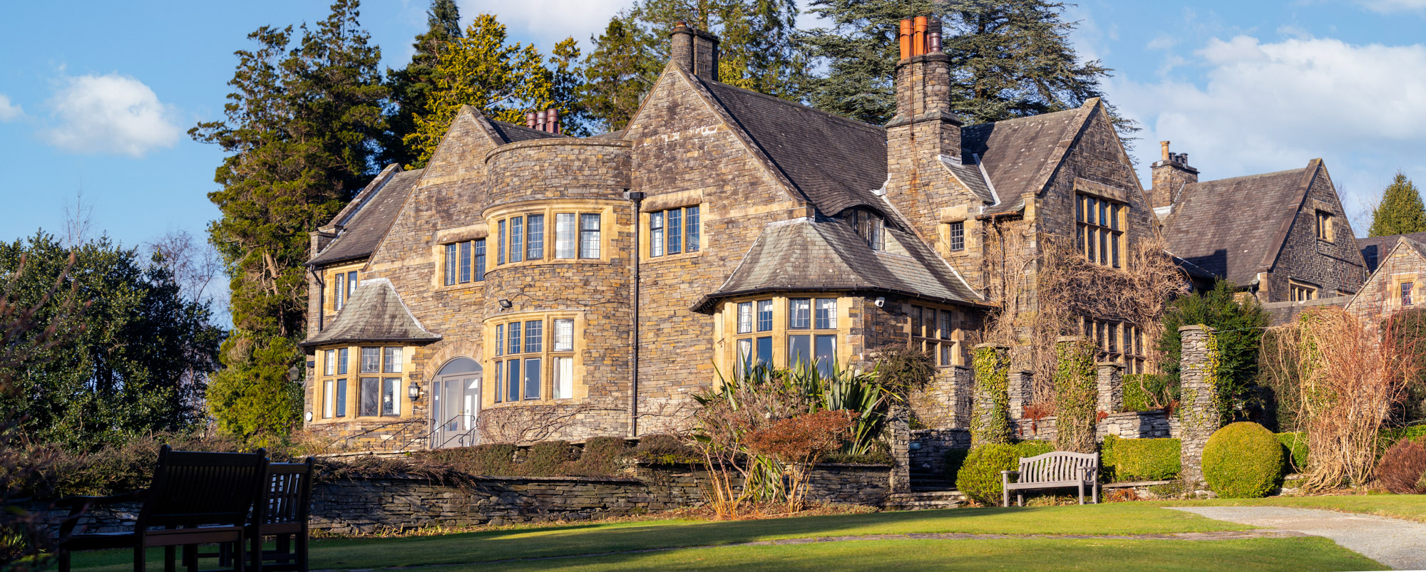 Panoramic exterior view of Cragwood Country House Hotel, Windermere — a two-storey Edwardian stone manor with mullioned bay windows, multiple chimneys and steeply pitched slate roofs, set in manicured gardens with topiary hedges and wooden benches, under a blue winter sky with light cloud.