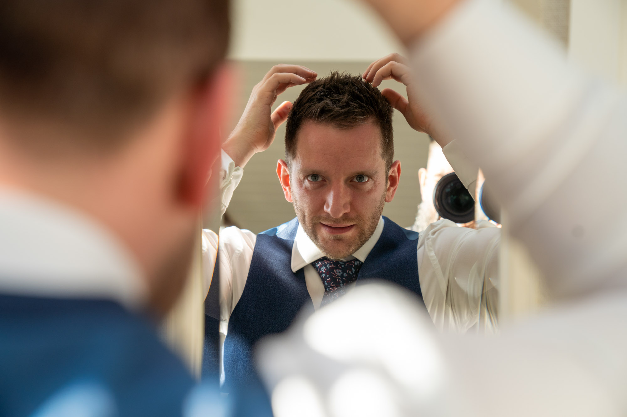 Groom in a navy three-piece suit and floral tie fixing his hair with both hands while looking into a mirror, photographed through a gap between two other guests in white shirts, at Cragwood Country House Hotel, Windermere.
