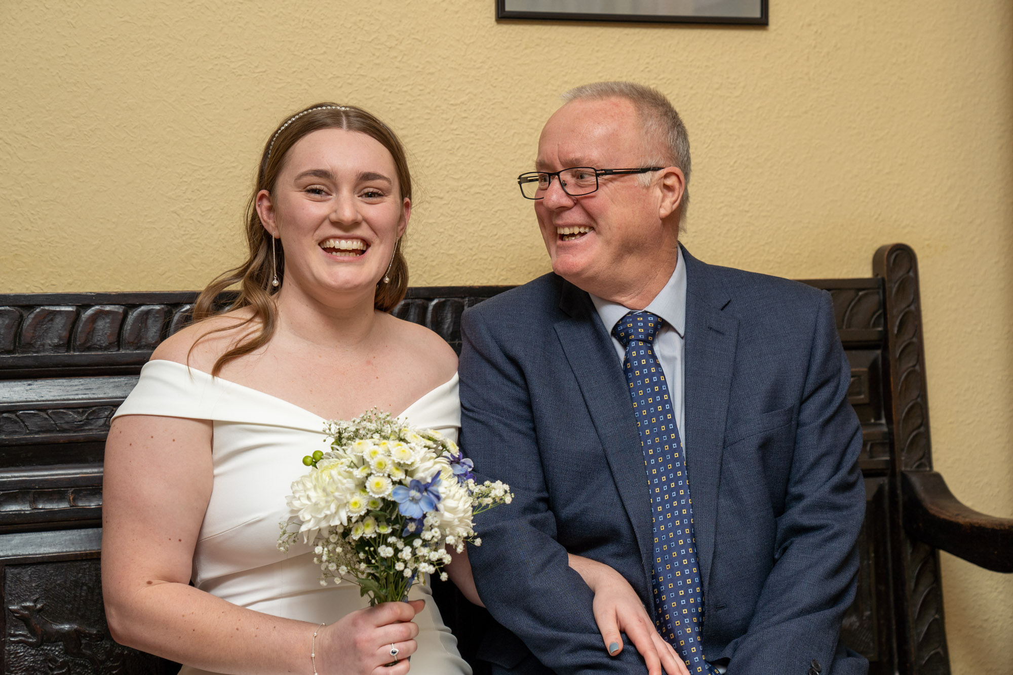 Bride in an off-the-shoulder white satin gown holding a small bouquet of white and blue flowers, laughing with an older man in a blue suit and glasses — likely her father — as they sit together on a carved dark wood bench inside Cragwood Country House Hotel.