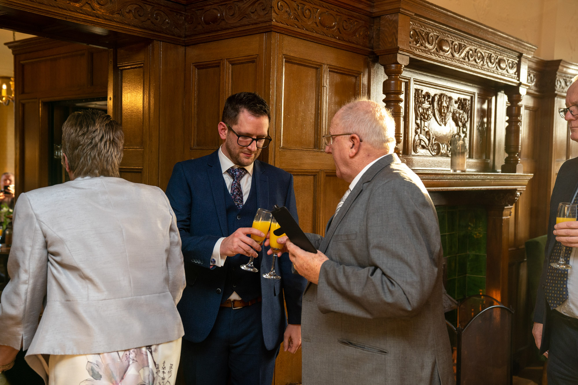 Groom in a navy three-piece suit holding a champagne flute of orange juice, standing by the ornately carved dark wood fireplace in the panelled interior of Cragwood Country House Hotel, talking with an older guest in a grey suit who is also holding a drink and showing him something on a tablet.