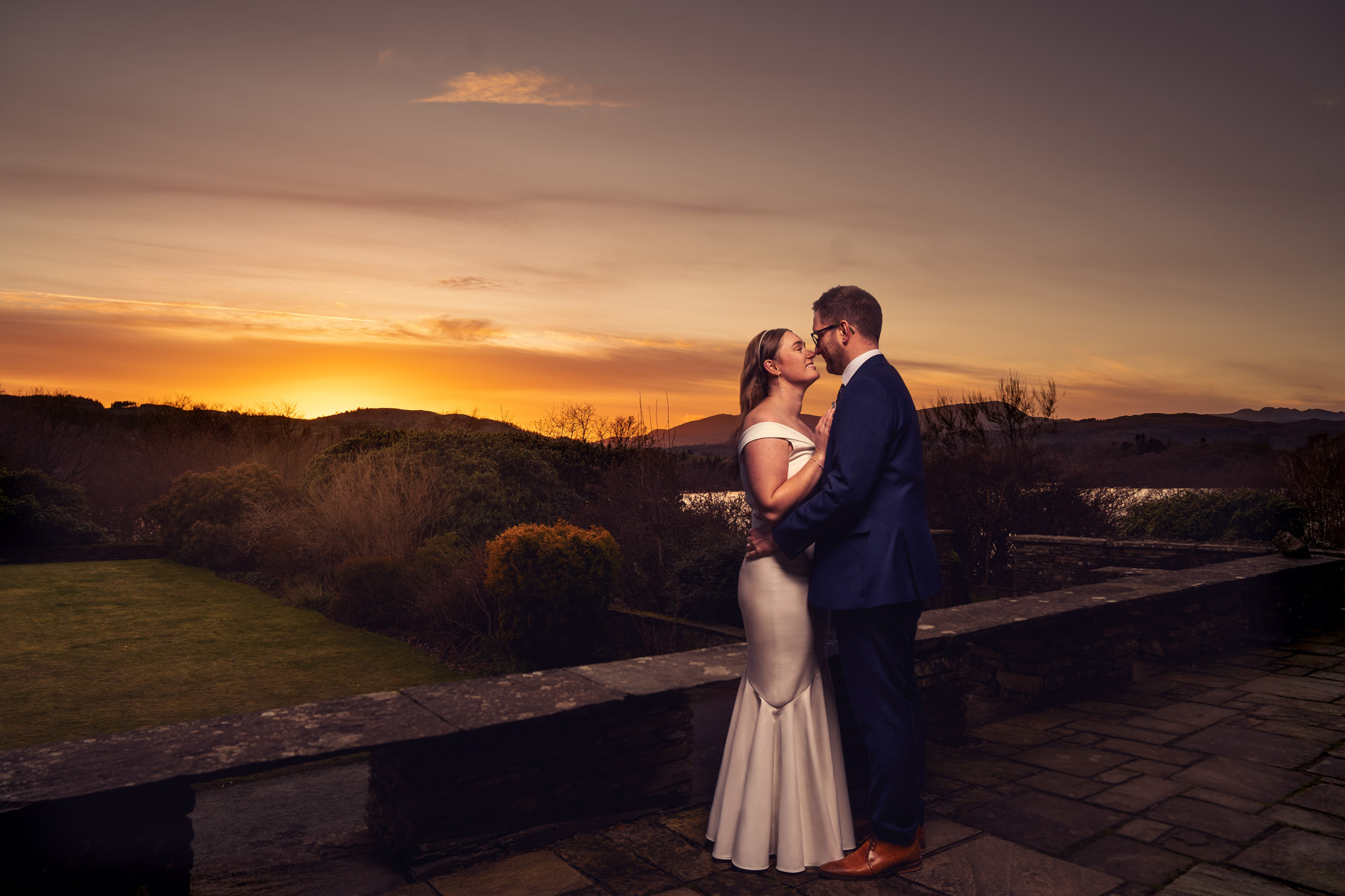 Bride and groom standing close together on the stone terrace at Cragwood Country House Hotel at dusk, looking at each other, with a vivid deep orange and amber sunset over the Lake District fells and a glimpse of Lake Windermere in the garden below.