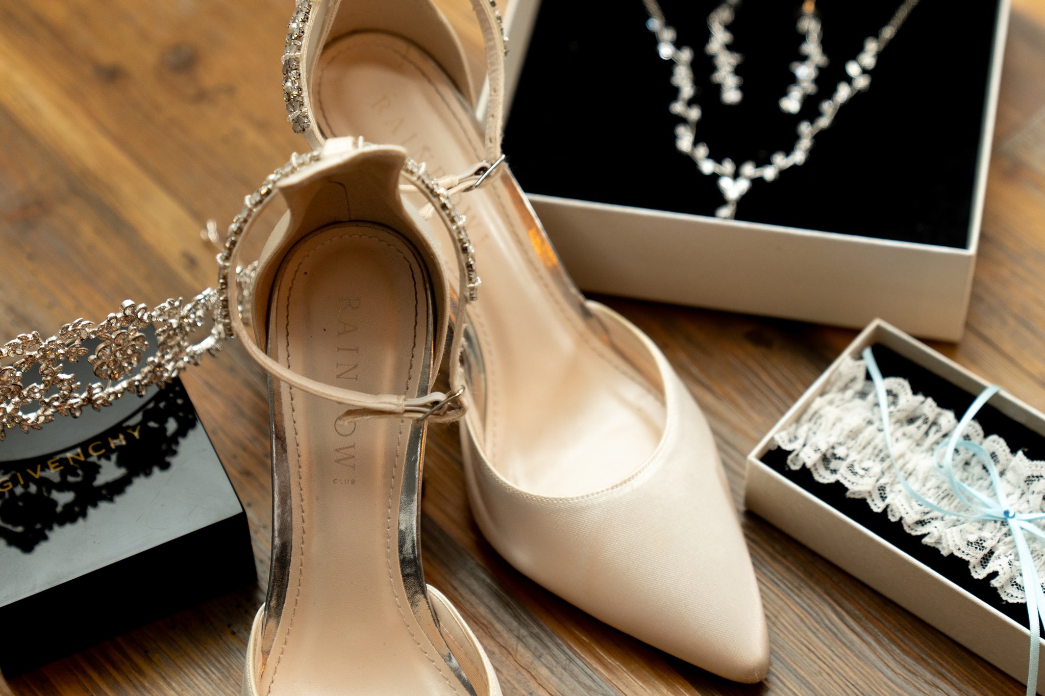 Flat-lay of bridal accessories on a wooden surface: ivory satin pointed-toe heels with crystal ankle straps, a white lace garter with blue ribbon in a box, a diamond necklace in a box, and a Givenchy perfume box to the left