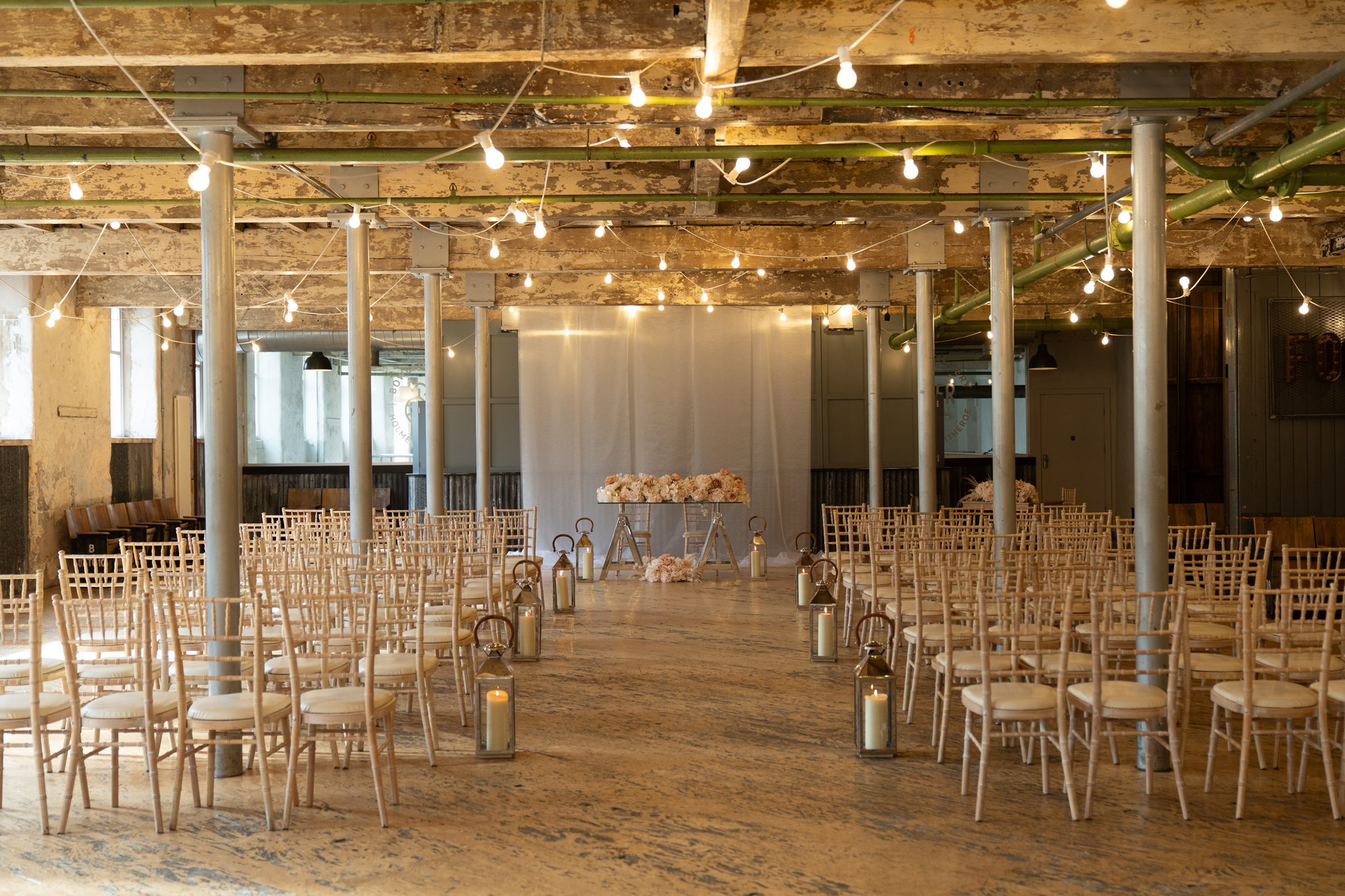 Holmes Mill ceremony room set for a wedding, wide shot showing rows of natural wood chiavari chairs with cream cushions, candle lanterns along the central aisle, festoon lights strung across the exposed industrial ceiling with green painted pipes and aged concrete columns, a draped white fabric backdrop and blush floral arrangement at the far end