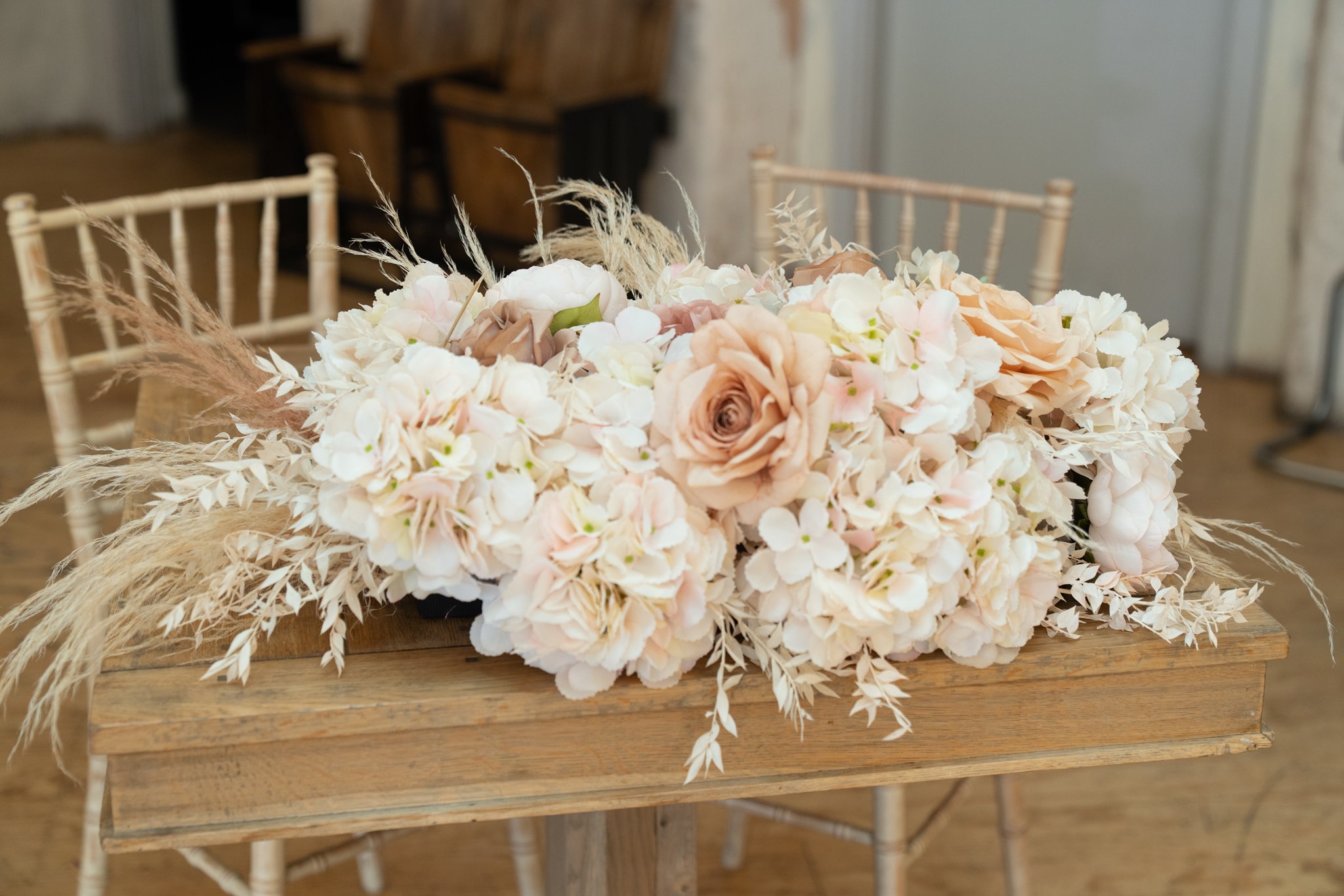 Close-up of a wedding ceremony floral arrangement on a rustic wooden trestle: blush and cream hydrangeas, peach roses and dried pampas grass spilling over the sides, chiavari chairs and white fabric backdrop softly blurred behind