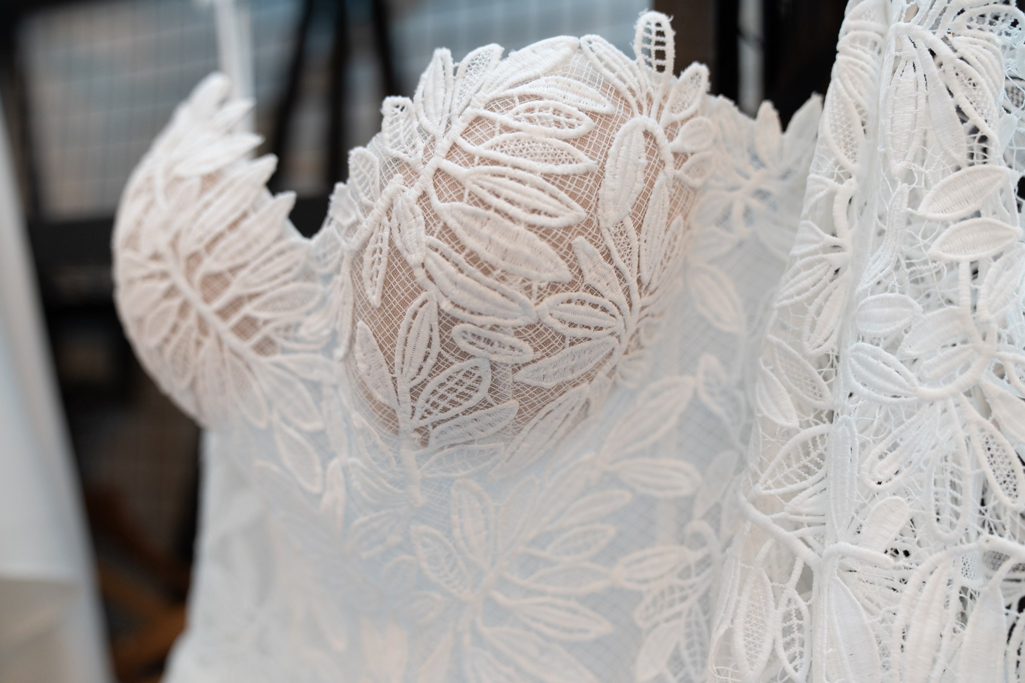Close-up detail of a white lace wedding dress bodice on a hanger, showing intricate leaf and petal appliqué lace on a sheer nude base, blurred background