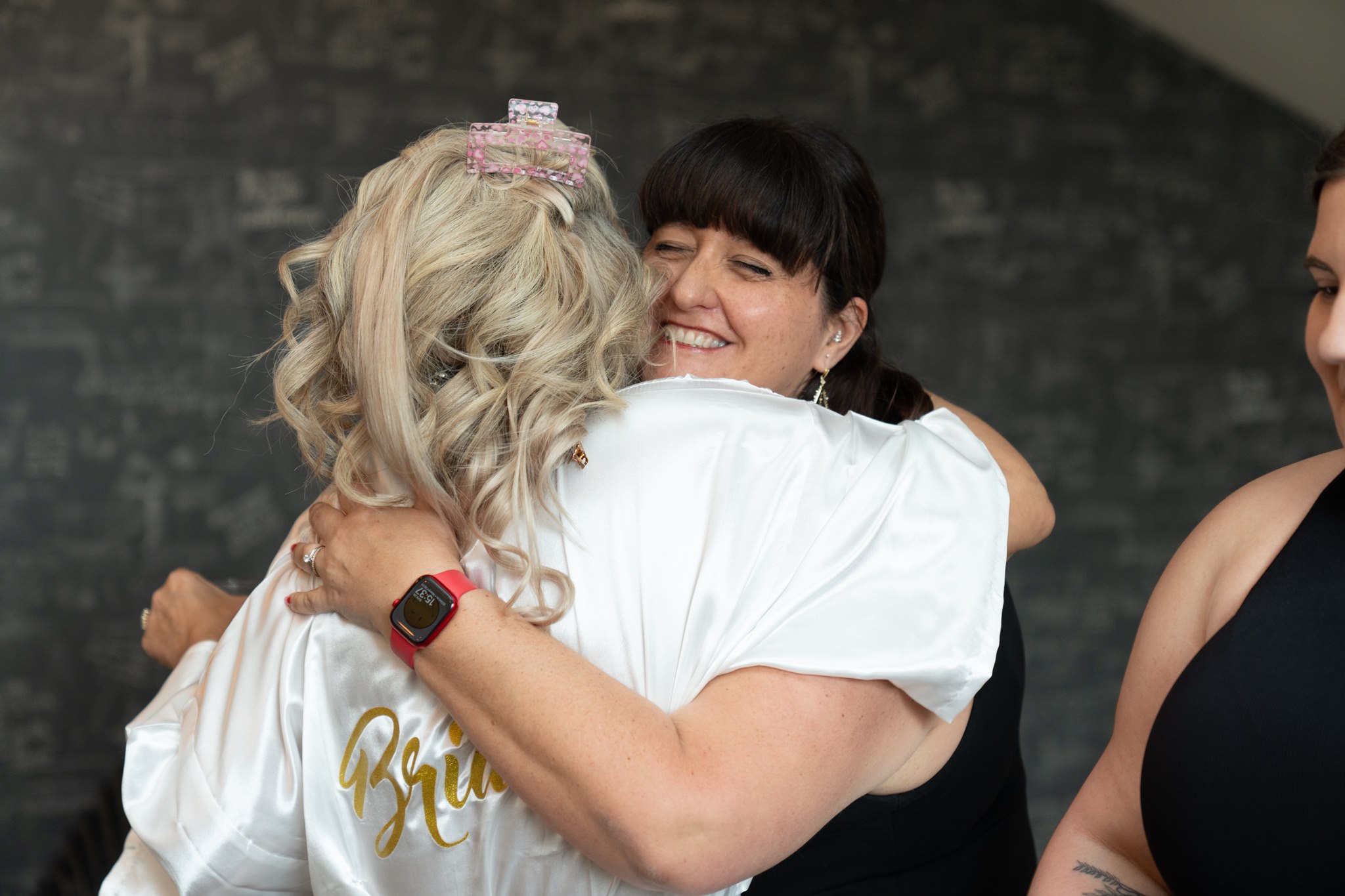 Bride in white satin robe hugging a dark-haired woman in a black top who is smiling broadly at the camera, against a dark textured wall, the back of the robe visible with gold Bride lettering