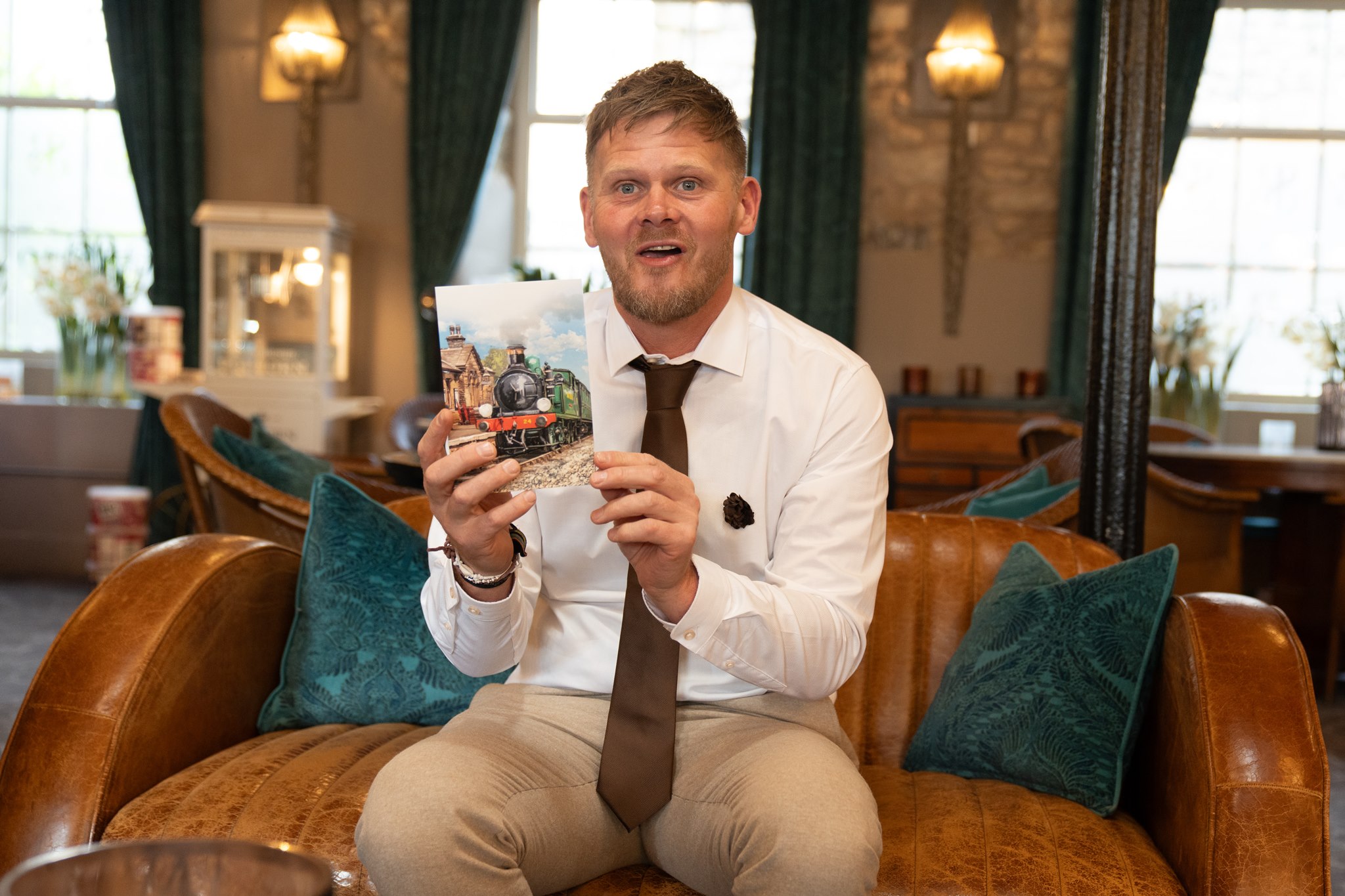 Groom in white shirt, brown tie and beige suit jacket sitting on a brown leather sofa holding up a wedding card with a steam train illustration, looking surprised and delighted, green velvet cushions and ornate hotel lounge behind him