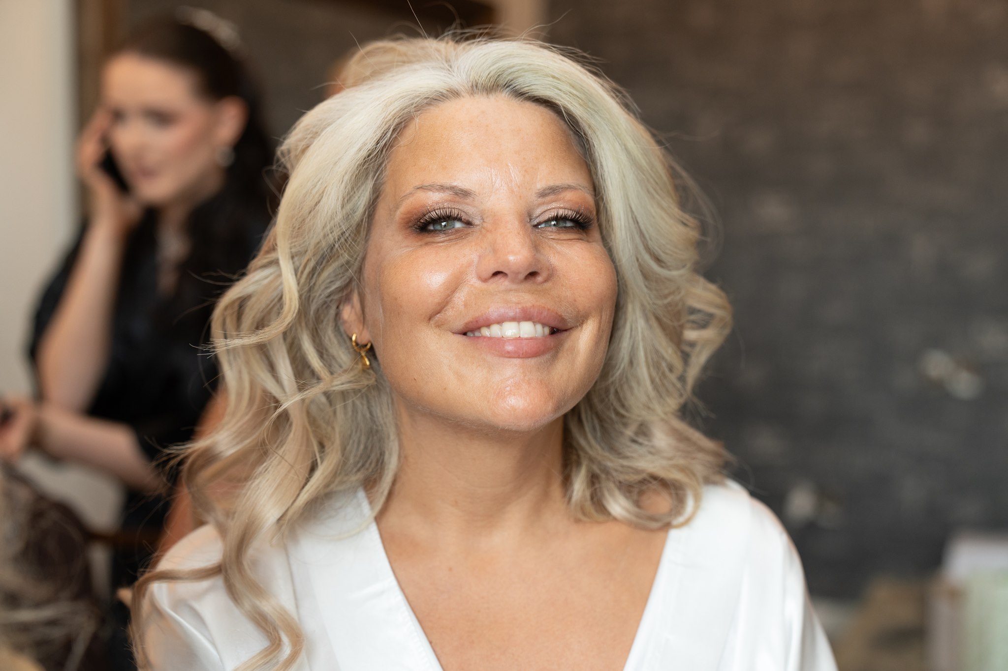 Bride in white satin robe smiling warmly at camera, silver blonde wavy hair down, gold hoop earrings, makeup complete, a stylist blurred on the phone in the background against a dark stone wall