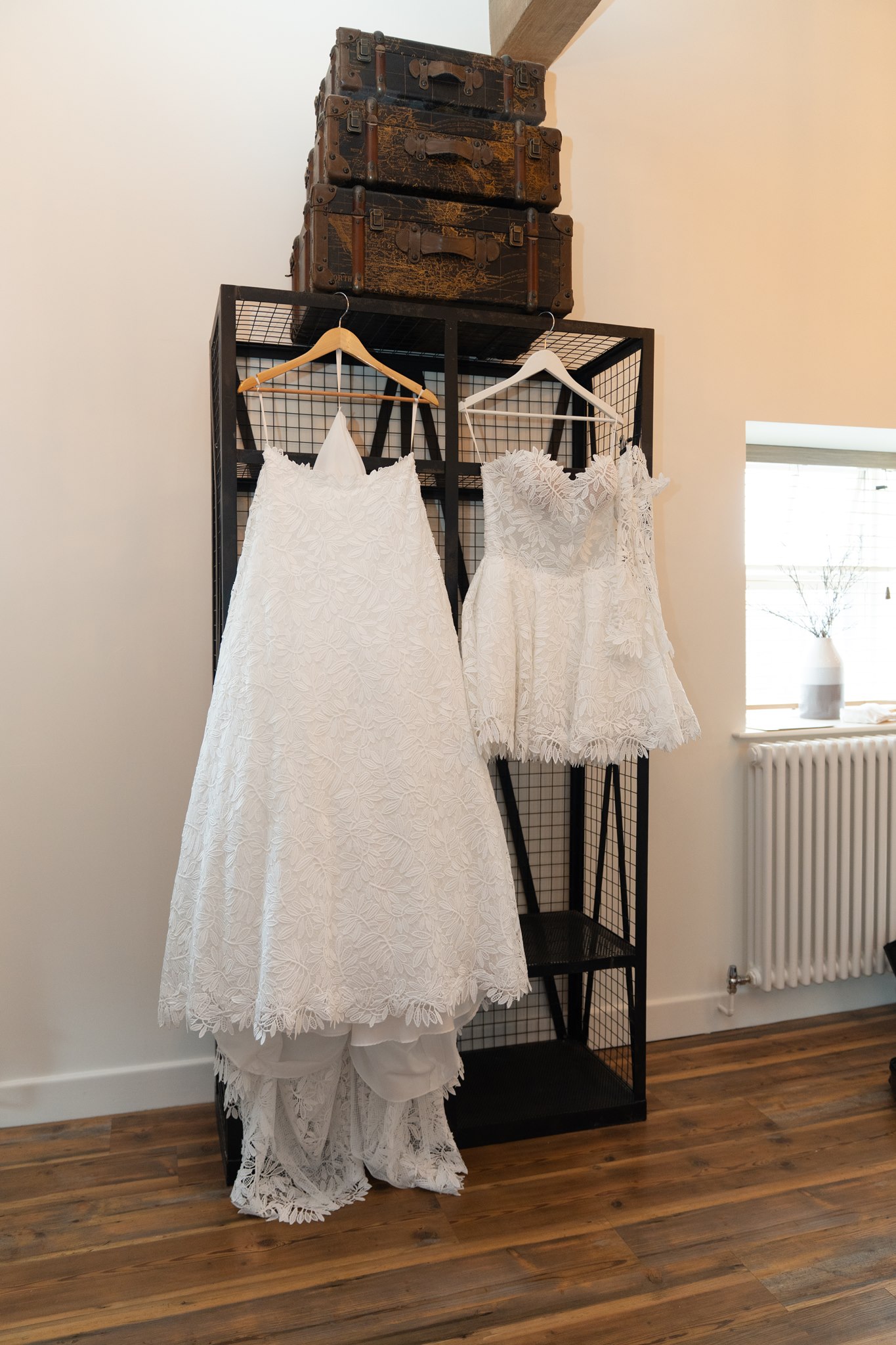 Two white lace wedding dresses hanging from a black metal frame storage unit with vintage stacked trunks above, one full-length gown and one shorter mini dress, wooden floor and neutral walls