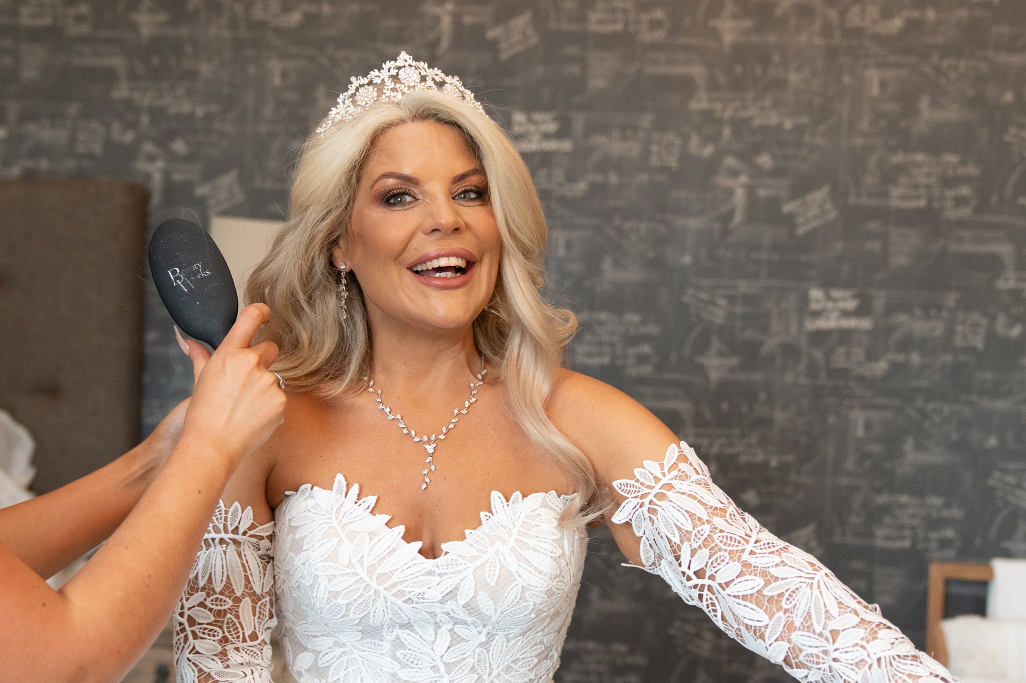 Bride in a white lace leaf-appliqué wedding dress and crystal tiara, laughing widely as a stylist brushes her silver blonde hair, diamond necklace and drop earrings on, dark chalk-covered feature wall behind her
