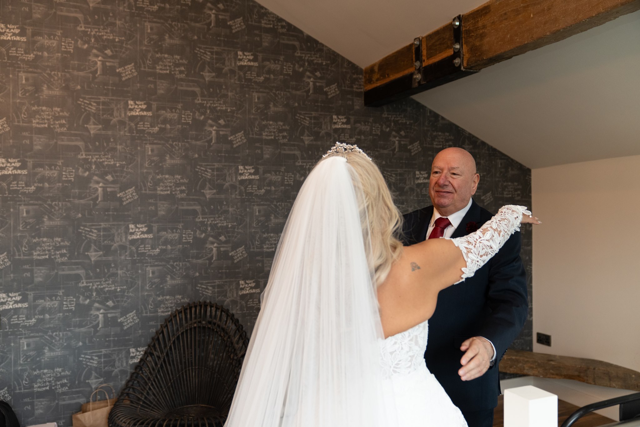 Bride in lace wedding dress with veil and crystal tiara facing an older man in a navy suit with red tie who looks at her with a quiet proud smile, against a dark chalk-covered wall with repeated motivational text at Holmes Mill Clitheroe