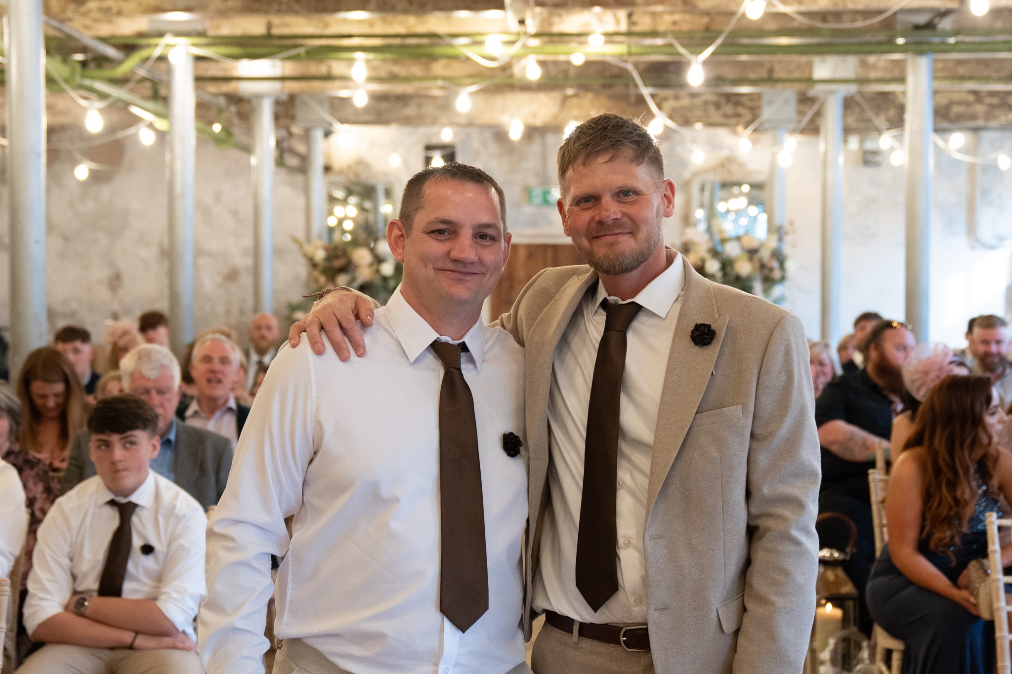 Groom in a beige suit with brown tie and dark floral buttonhole standing with his best man in a white shirt and matching brown tie, arm around each other, smiling at camera inside the Holmes Mill ceremony room with festoon lights and seated wedding guests behind them